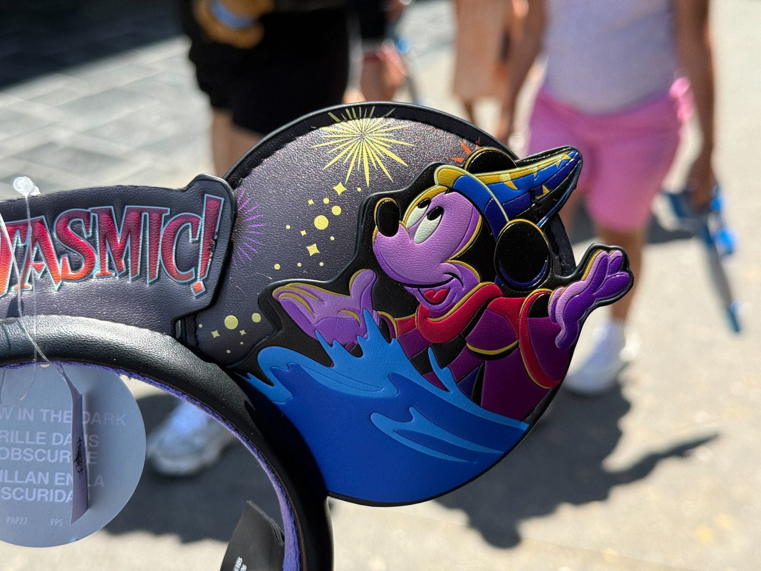 A Fantasmic Ear Headband with Sorcerer Mickey design, held up at Disneyland Resort with people in the background.
