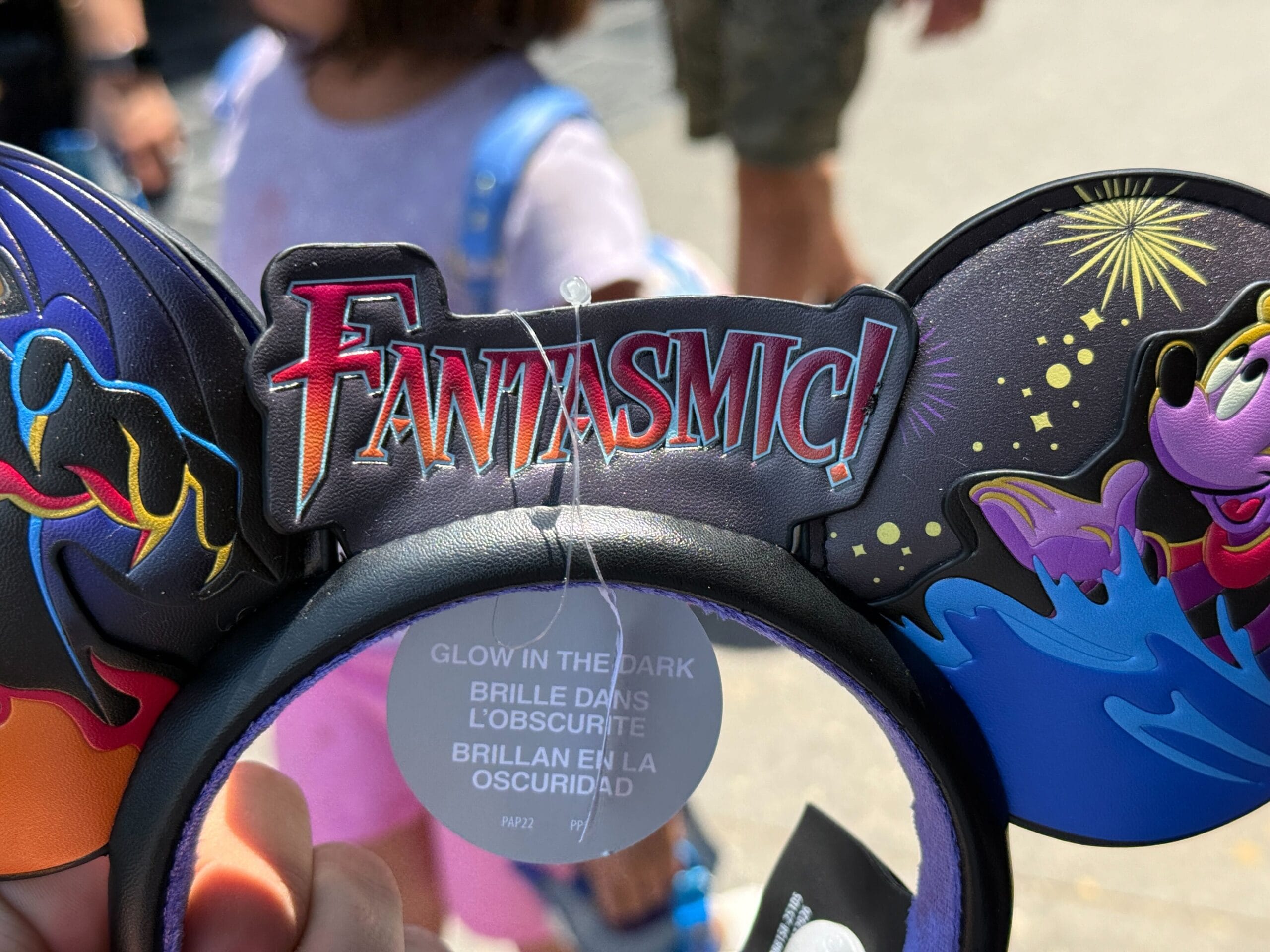 A hand holds a Fantasmic Ear Headband with "Fantasmic!" and glow-in-the-dark tag at Disneyland Resort, fireworks in back.