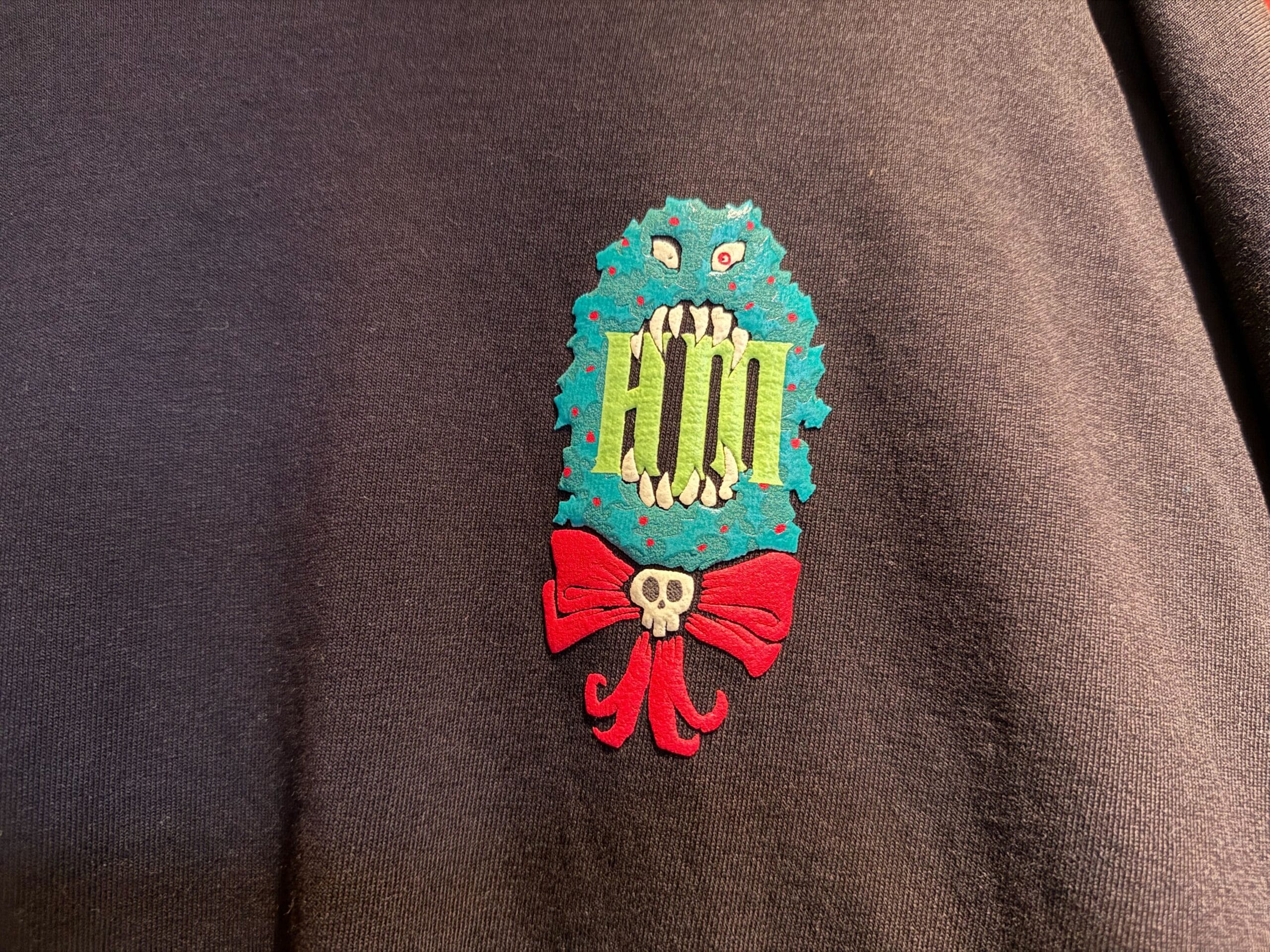 A blue monster face with sharp teeth, green "HM," and a red bow with a skull, inspired by Haunted Mansion Holiday.