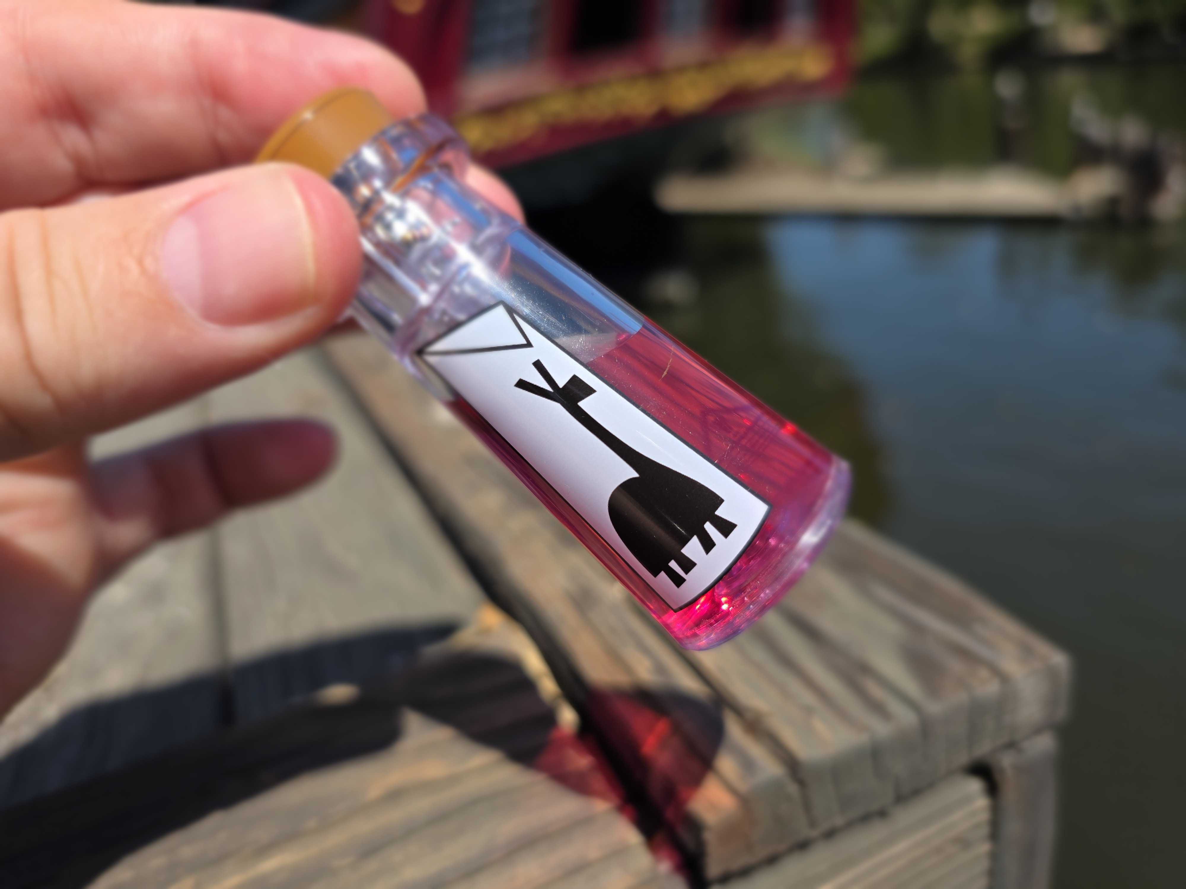 A hand holds Kuzco’s Poison Bottle, a vial with pink liquid and a giraffe label, near water at Disneyland.