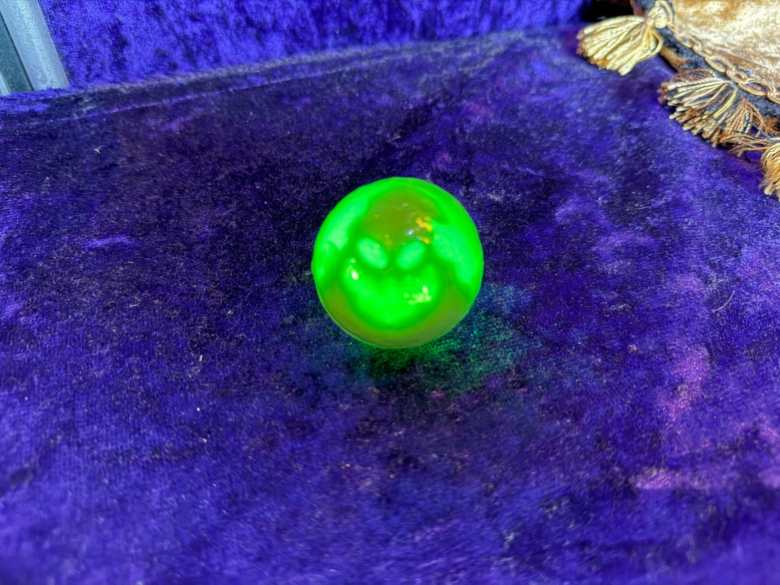 A shiny green ball sits on a purple velvet surface near a gold-trimmed fabric.