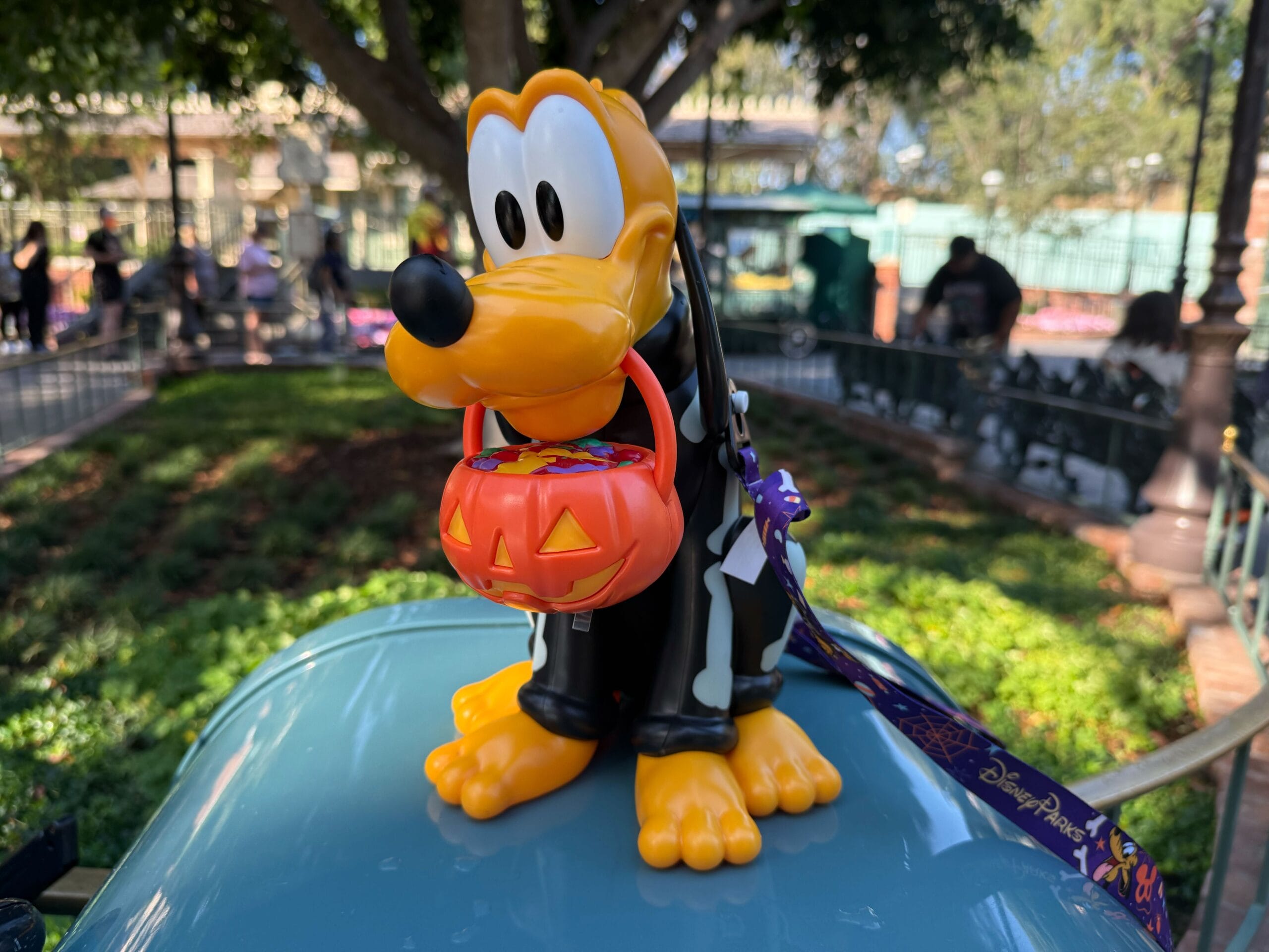 Pluto figurine dressed in a skeleton costume holds a pumpkin treat bucket, sitting on a blue surface outdoors.