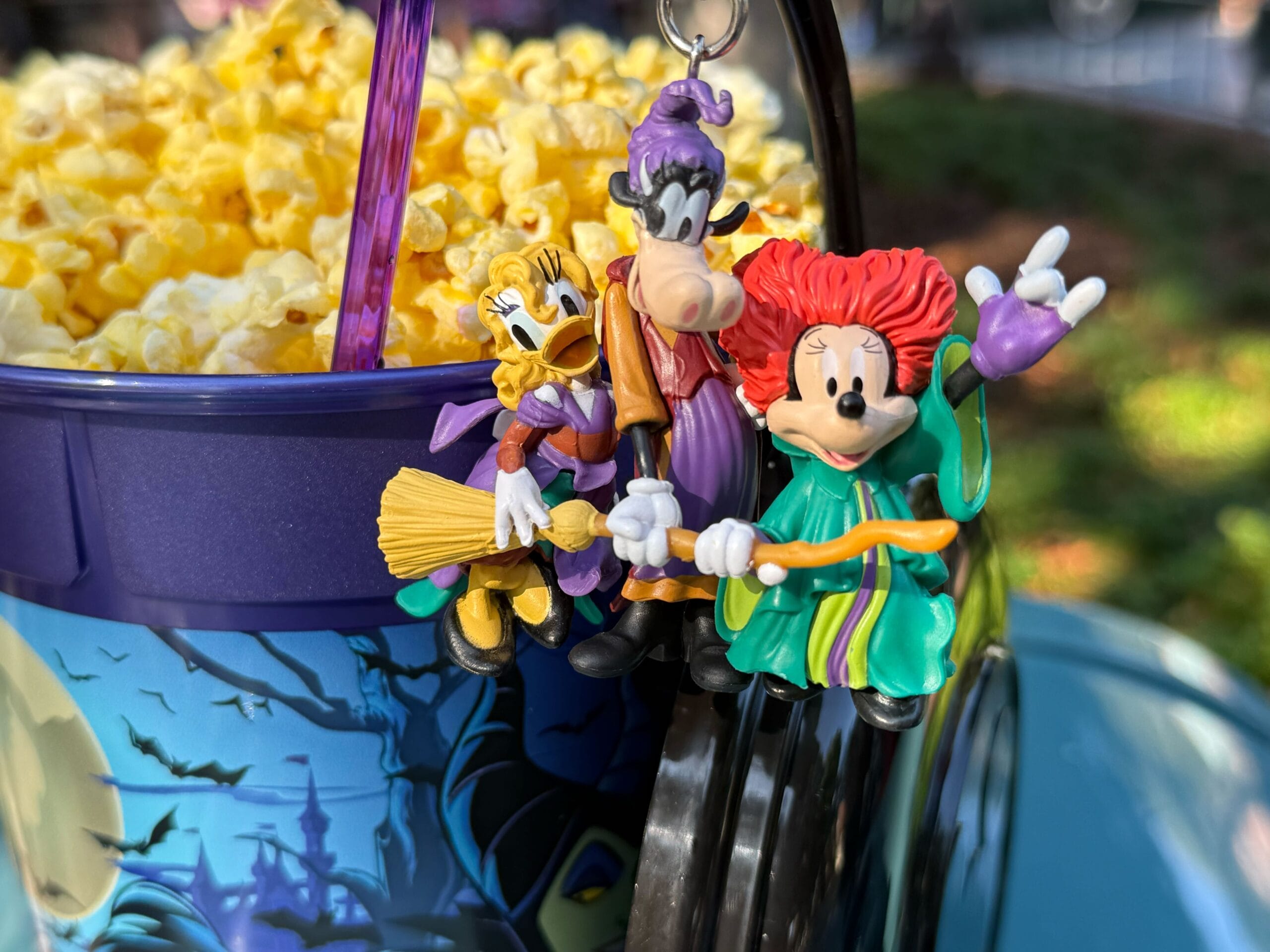 Three Disney characters in Halloween costumes dangle from a Disneyland popcorn bucket outdoors this Halloween season.