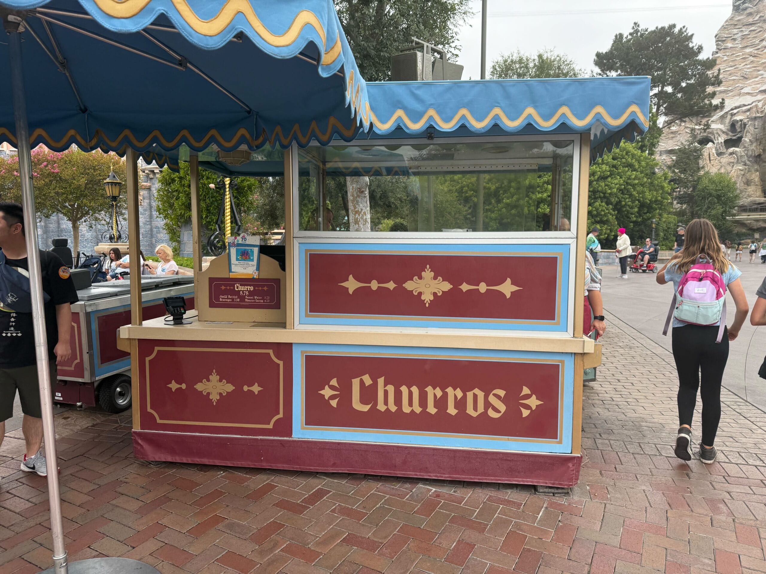 A red and gold churro stand with a blue canopy at an outdoor location, people walking nearby.