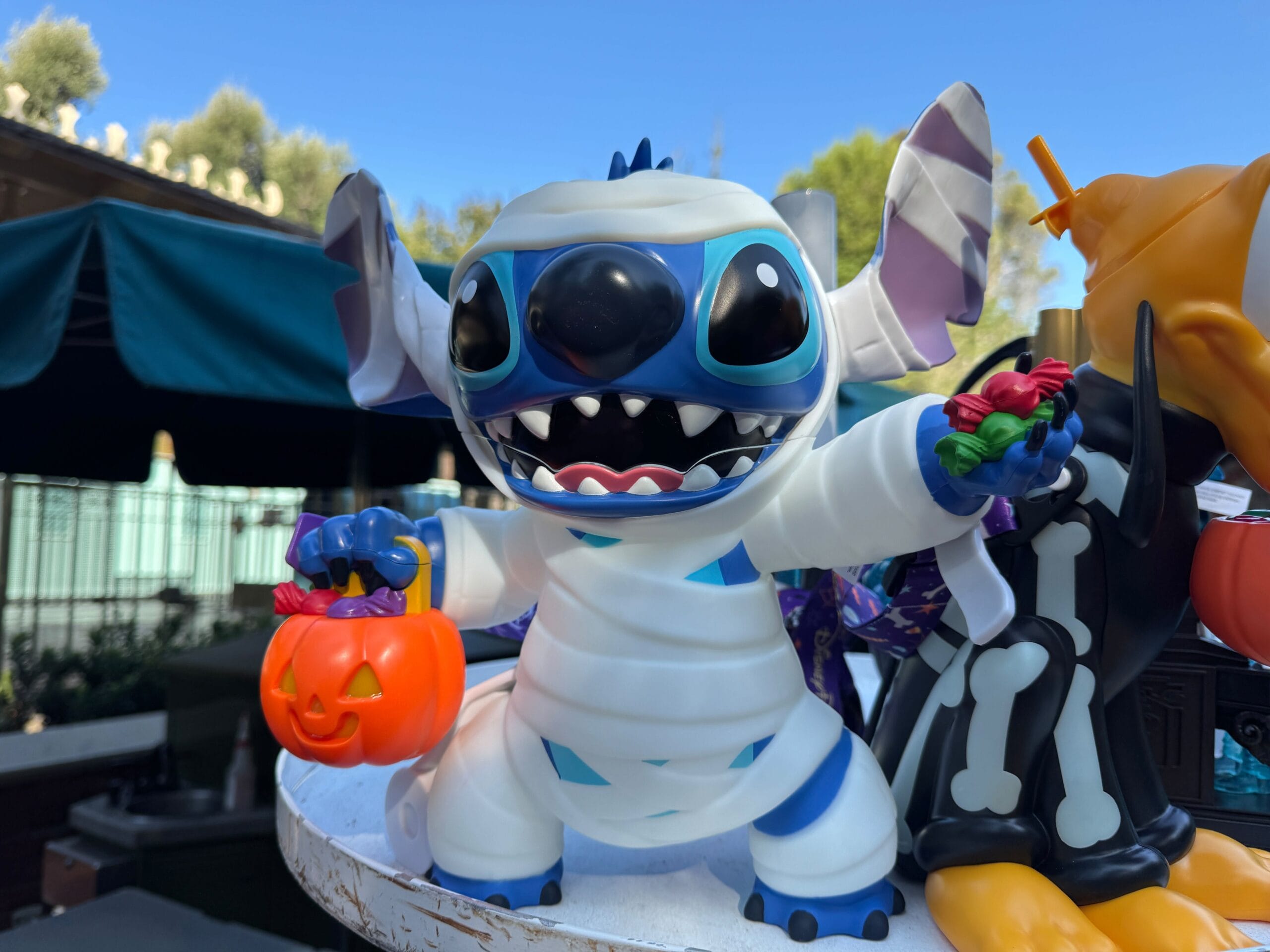 Stitch Mummy figurine holds a pumpkin bucket and candy, with another festive Halloween figure by his side.