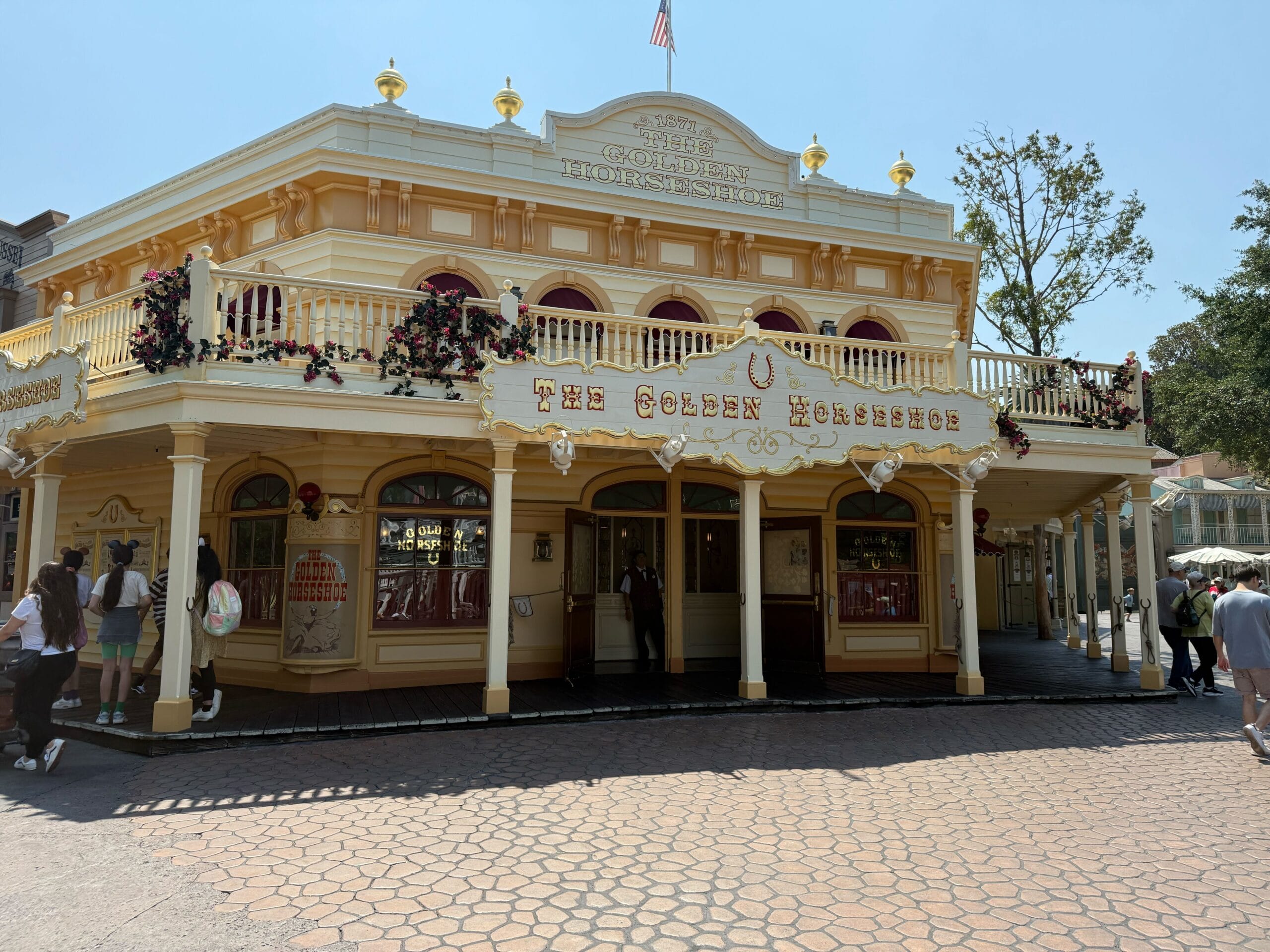 The Golden Horseshoe building at Disneyland, with yellow trim, balcony, and people walking nearby on a sunny day.