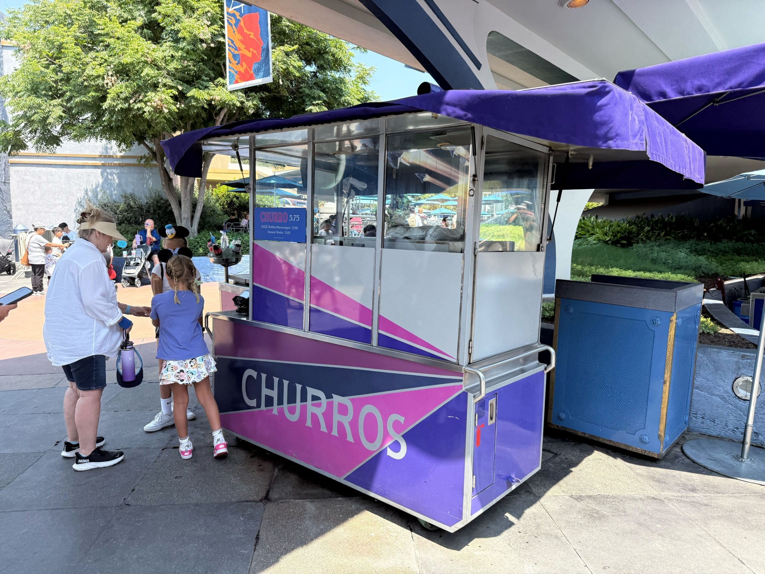 A purple churro cart with a canopy, serving two children and an adult in a sunny outdoor setting.