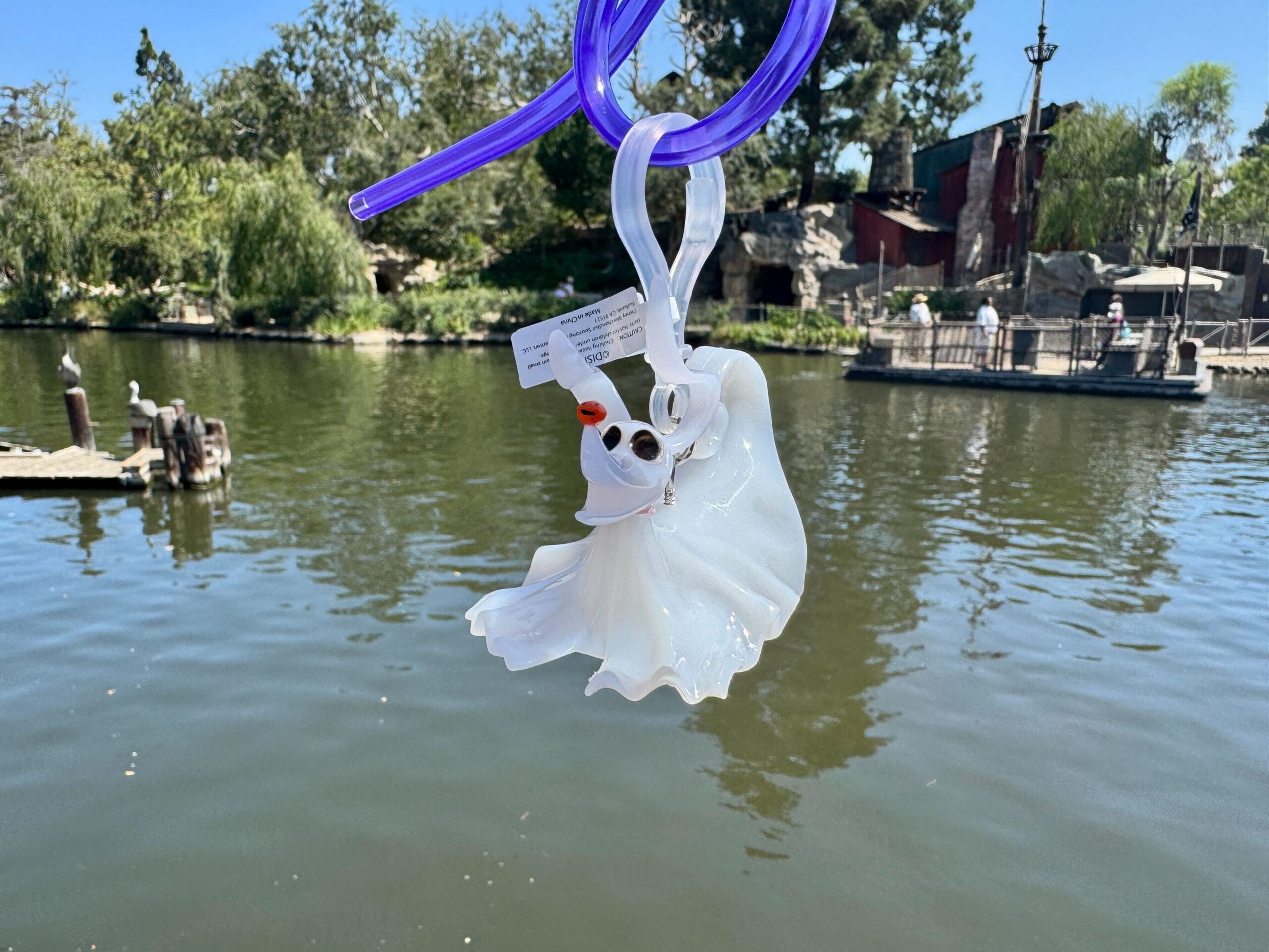 A white ghost-shaped drink holder with a red nose hangs over a lake, with trees and people in the background.
