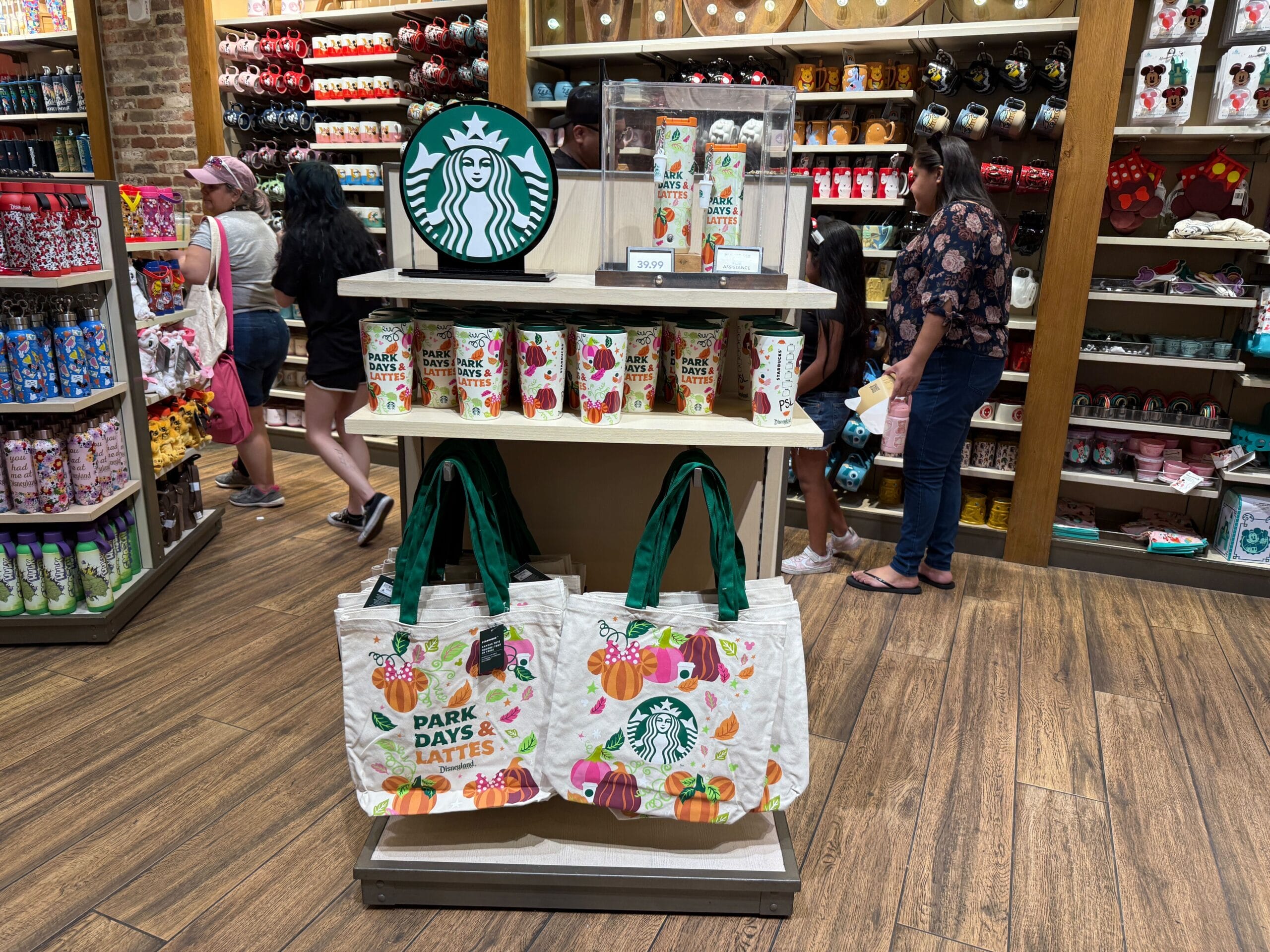 Starbucks merchandise display with tote bags, tumblers, and cups at Disneyland Resort as people shop in-store.