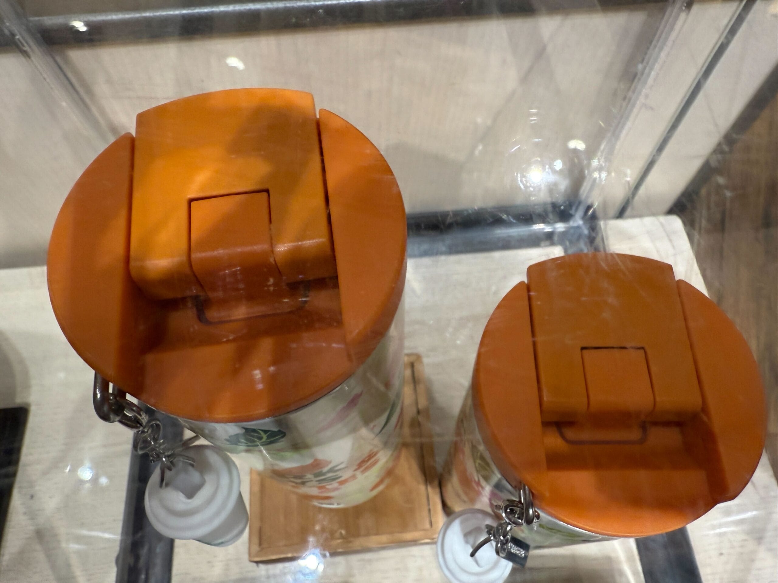 Two stainless steel travel mugs with orange lids, inspired by Starbucks Pumpkin Spice, sit behind a glass case.