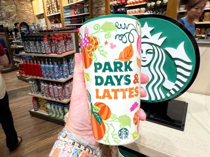 A hand holds a Starbucks Pumpkin Spice tumbler with pumpkins and "PARK DAYS & LATTES" text in a store.