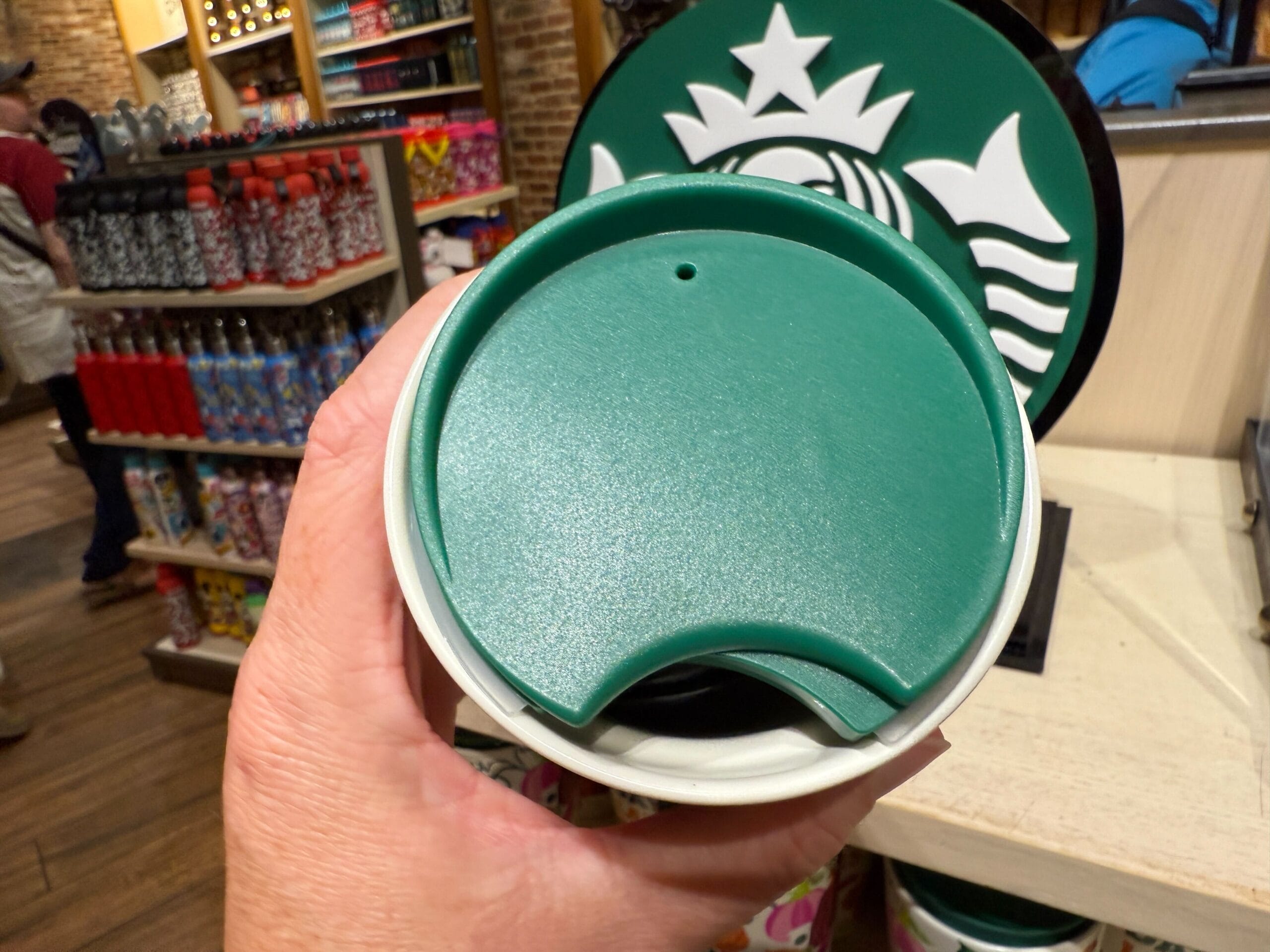 A hand holds a Starbucks Pumpkin Spice cup with a green lid inside a store lined with colorful bottles on shelves.