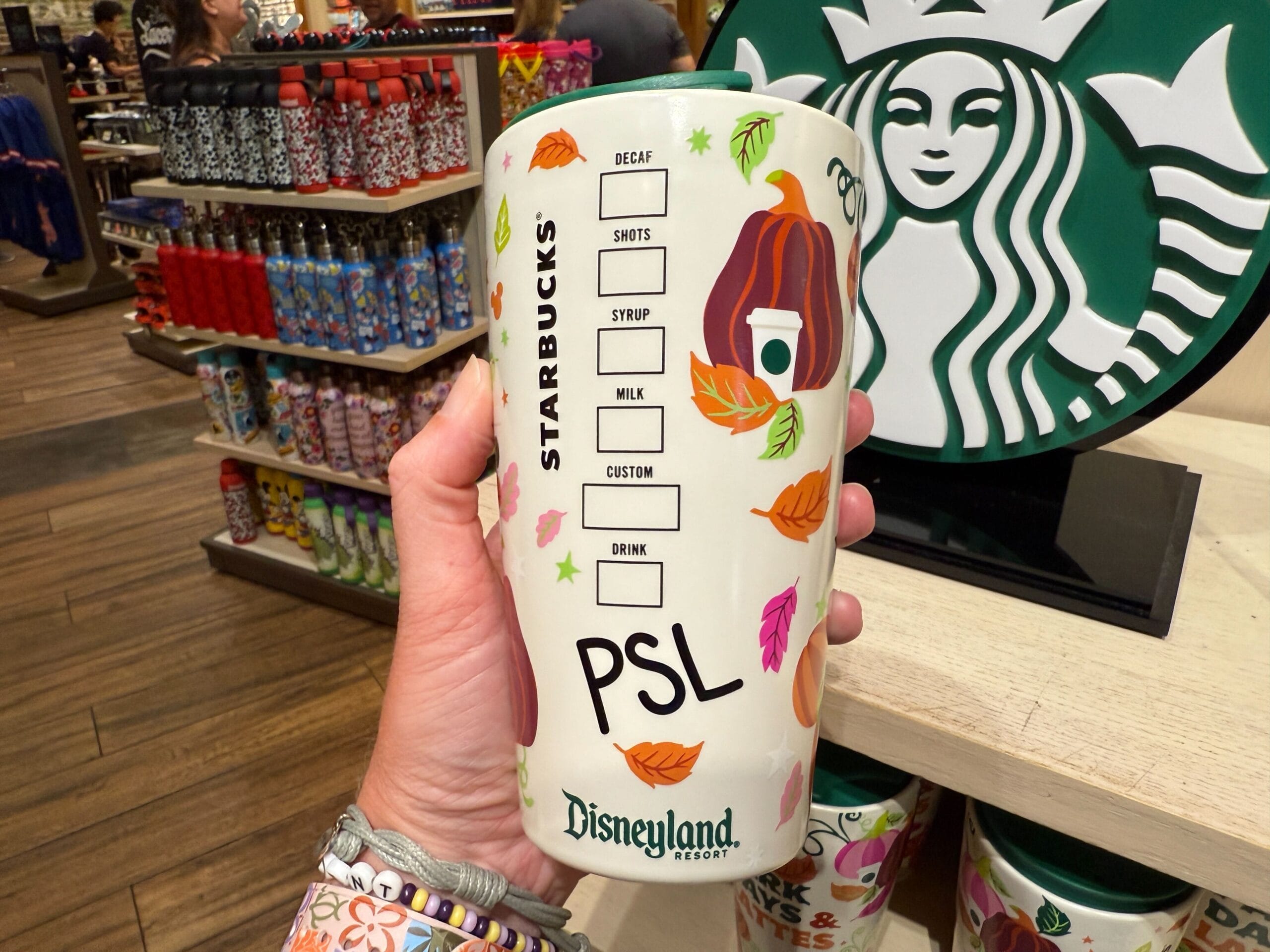 Hand holding a Starbucks Pumpkin Spice cup with fall-themed decor at a Disneyland Resort store.