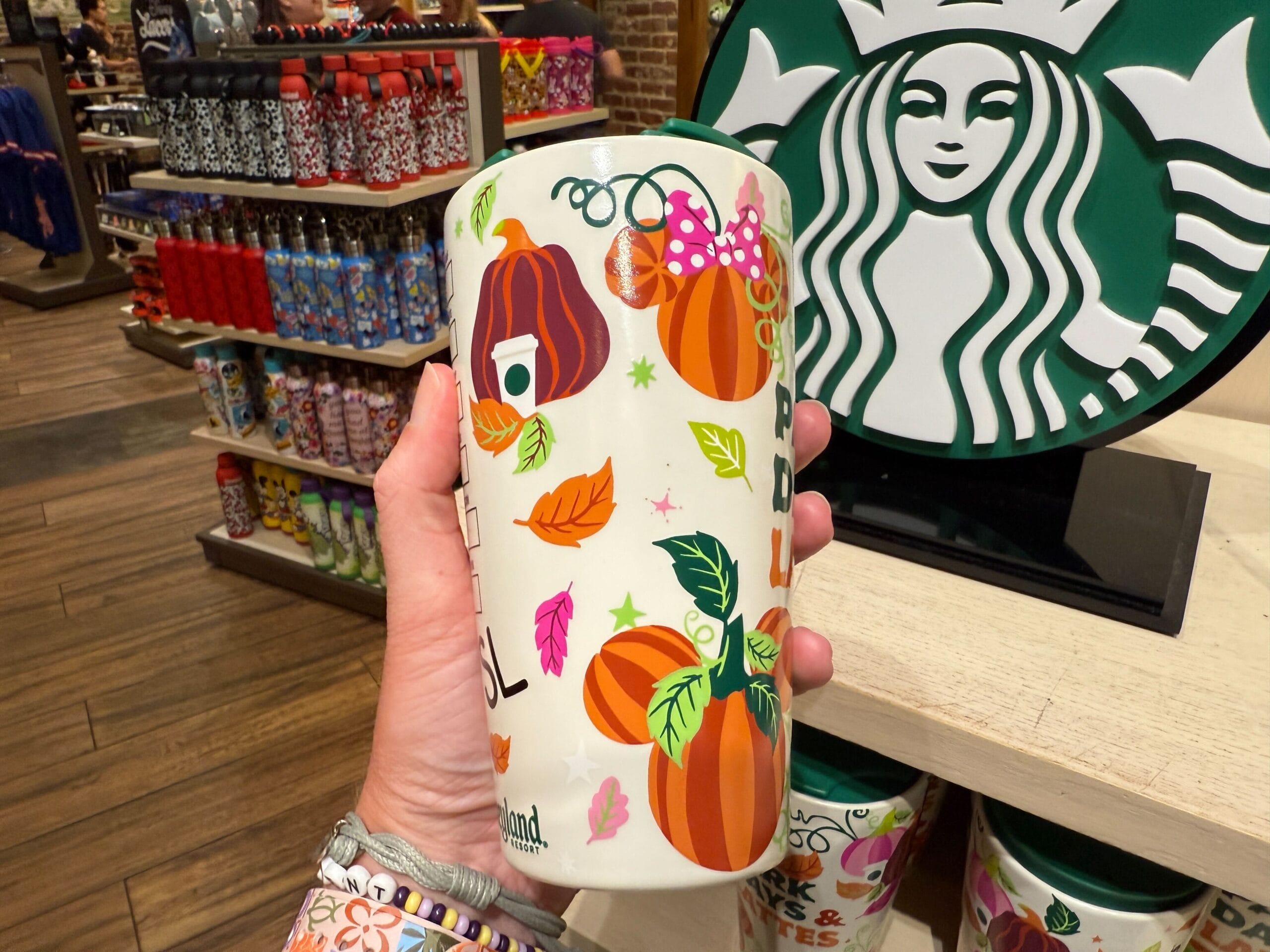 A hand holds a Starbucks Pumpkin Spice tumbler with fall pumpkin and Minnie Mouse bow designs in a store.