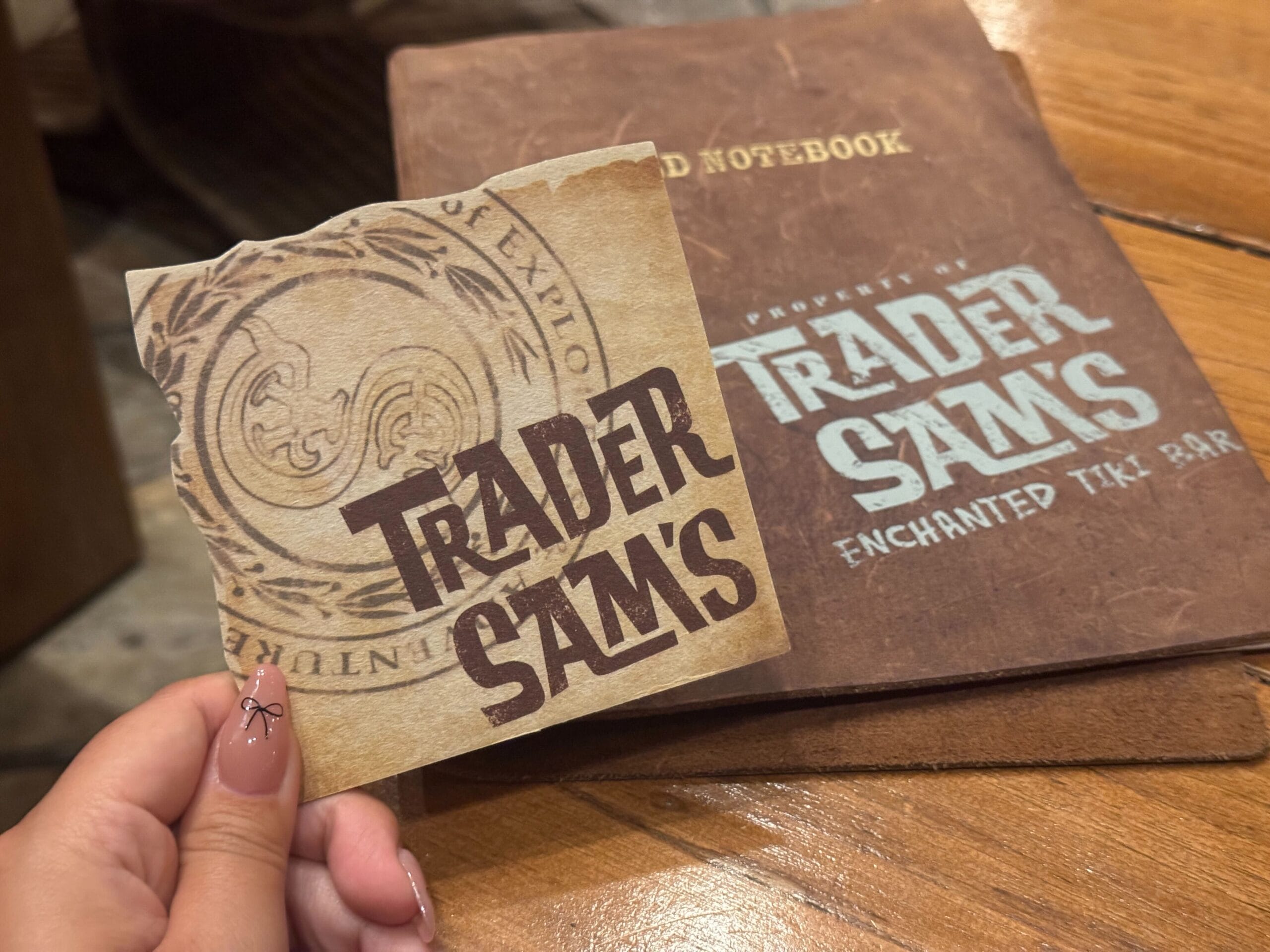 A hand showcases a New Arrival: Trader Sam's Coasters at Disneyland, in front of a brown Tiki Bar notebook on the table.