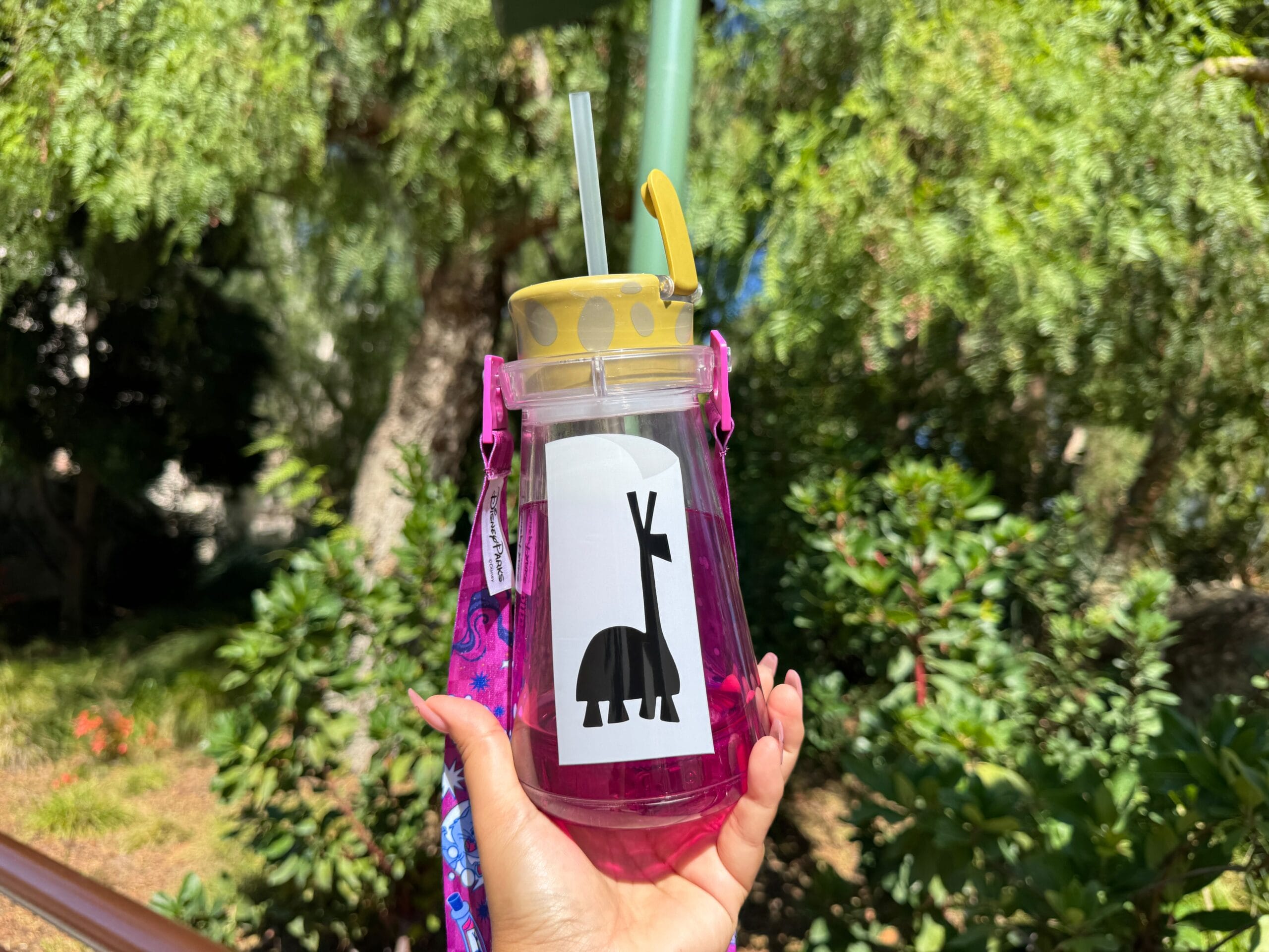 A hand holds a pink Light Up Kuzco's Poison Sipper with a straw and llama sticker, outdoors at sunny Disneyland.