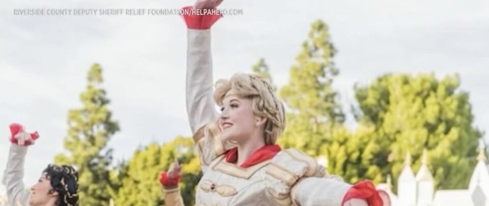 A performer in a gold and white costume raises one arm during a Disneyland parade, with trees and buildings in the background.