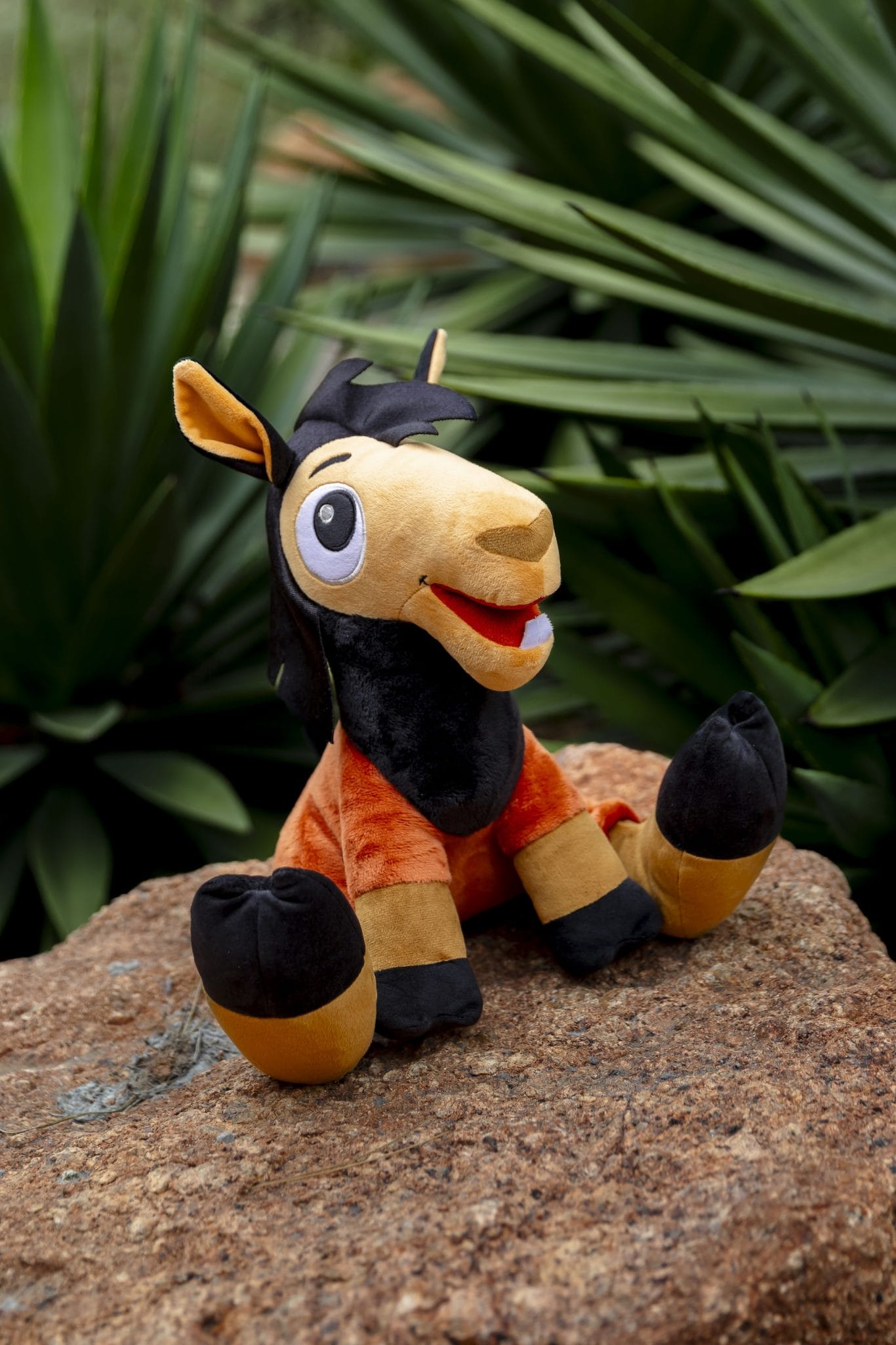 A plush Kuzco llama with orange and black fur sits on a rock with green plants in the background.