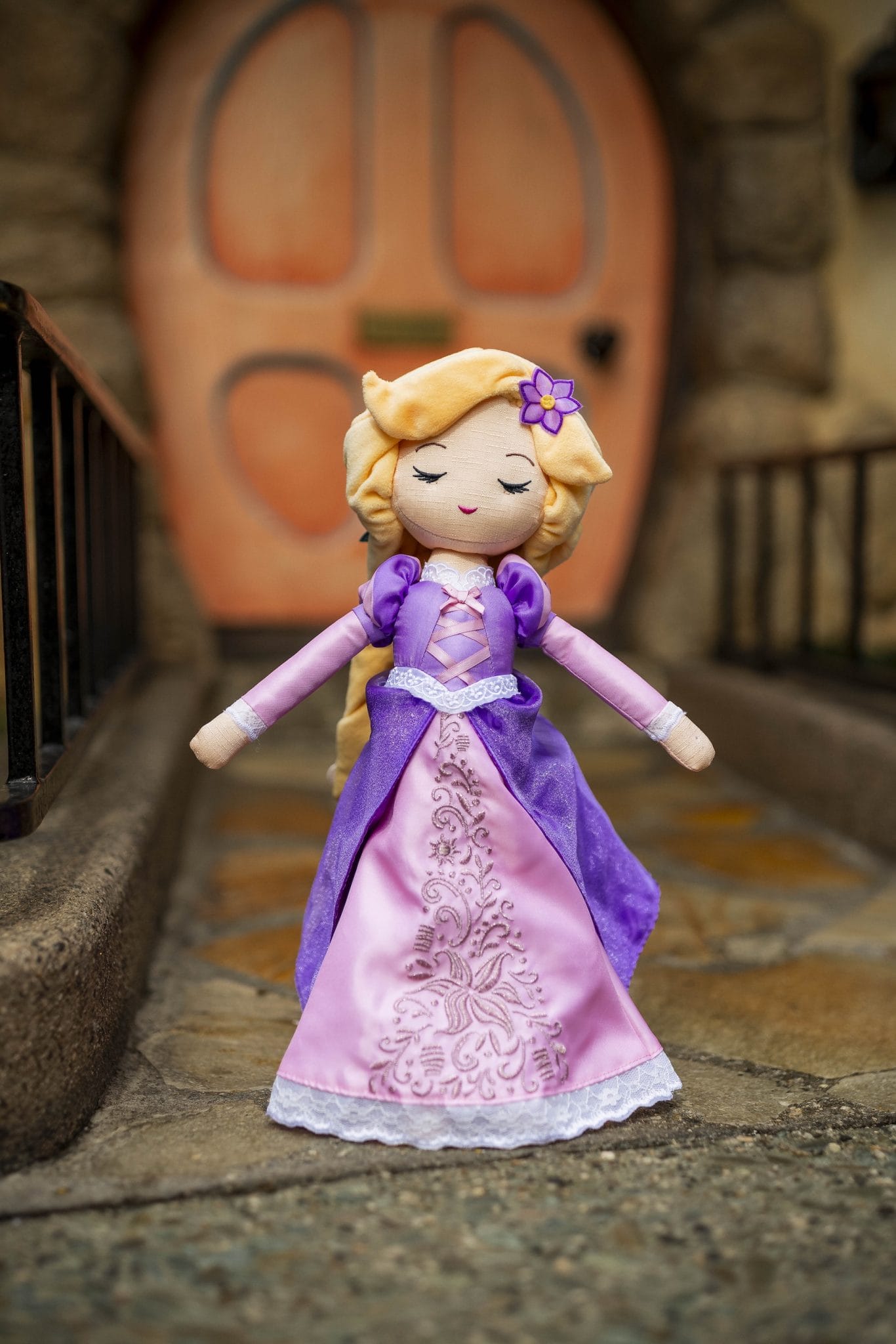 A Rapunzel doll in a purple and pink dress stands beside Disney Shimmer Wishables on a stone path by a wooden door.