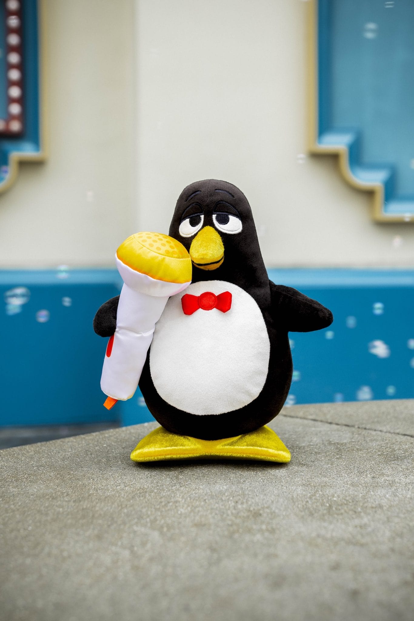 A black and white Wheezy plush toy with a red bow tie holds a yellow Mini MixIts microphone outdoors.