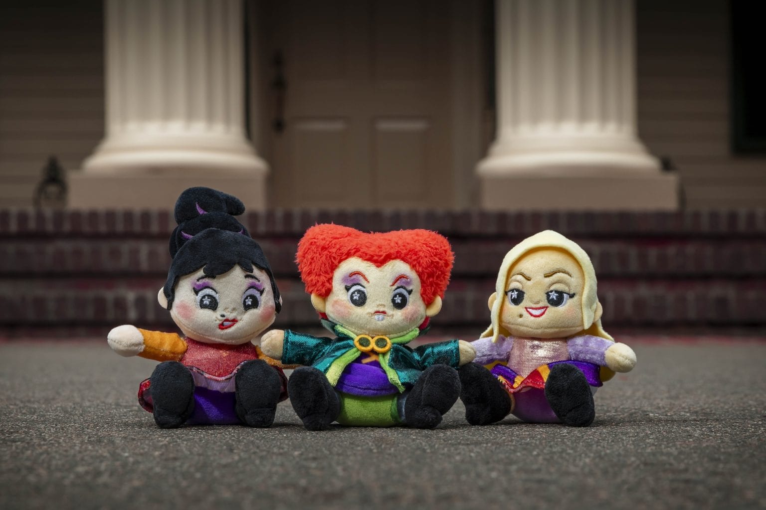 Three plush shimmer wishables dolls dressed as witches sit side by side on pavement, columns and a door behind them.