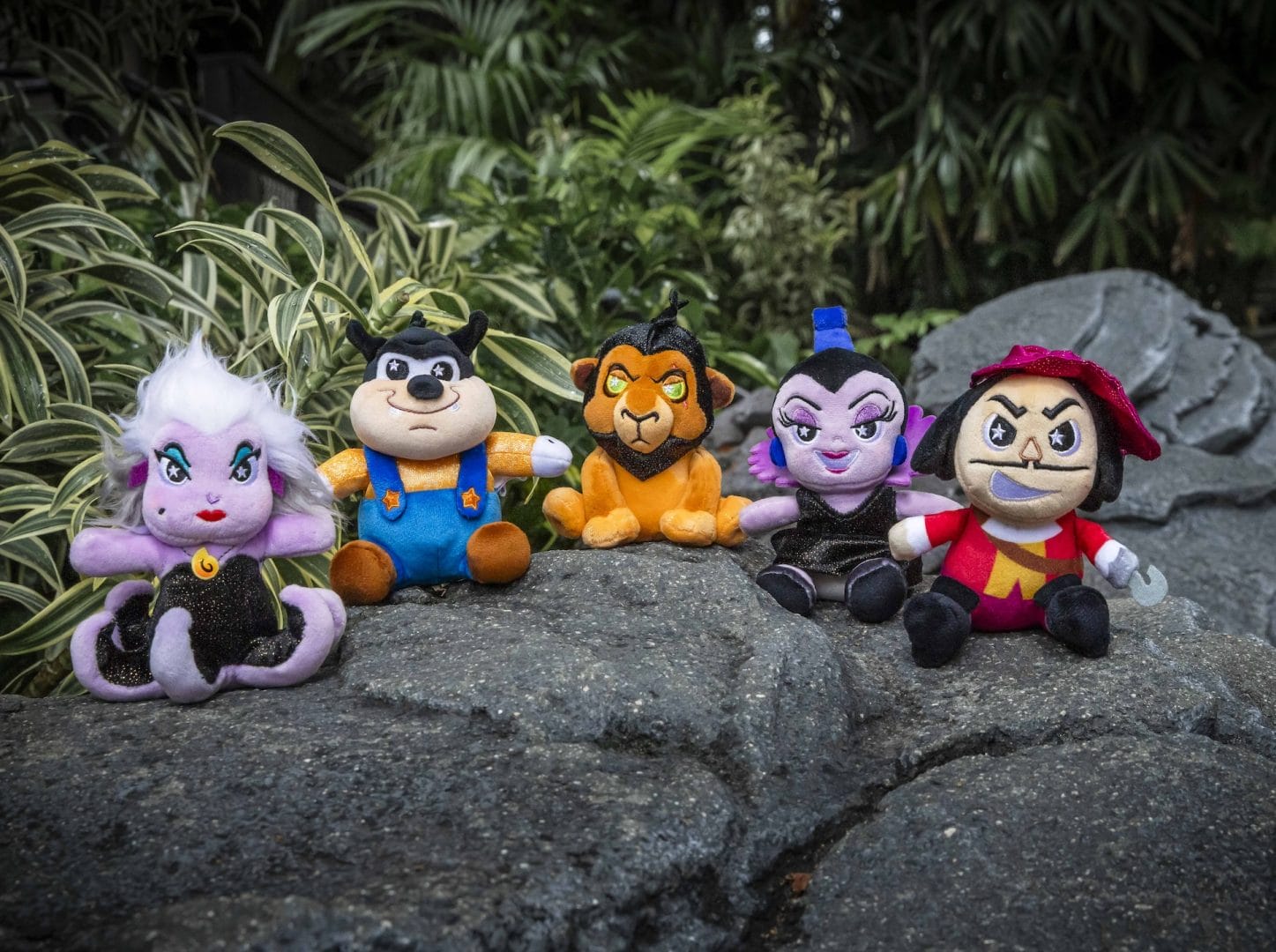 Five Disney villain plush toys, including Squishmallows, sit on a rock outdoors with green plants in the background.