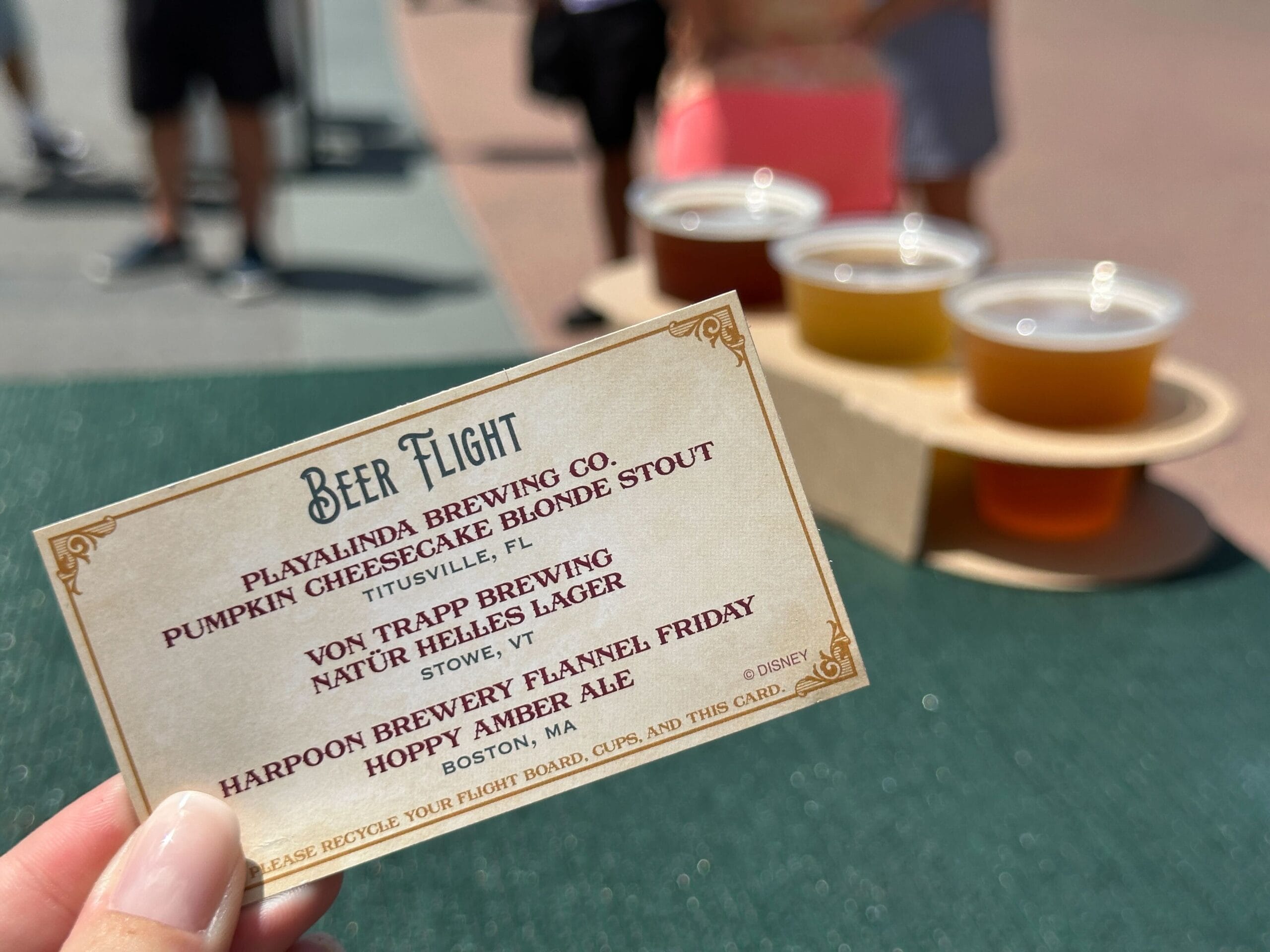 A hand holds a beer flight menu with four beers on a tray in the blurred Food & Wine outdoor background.