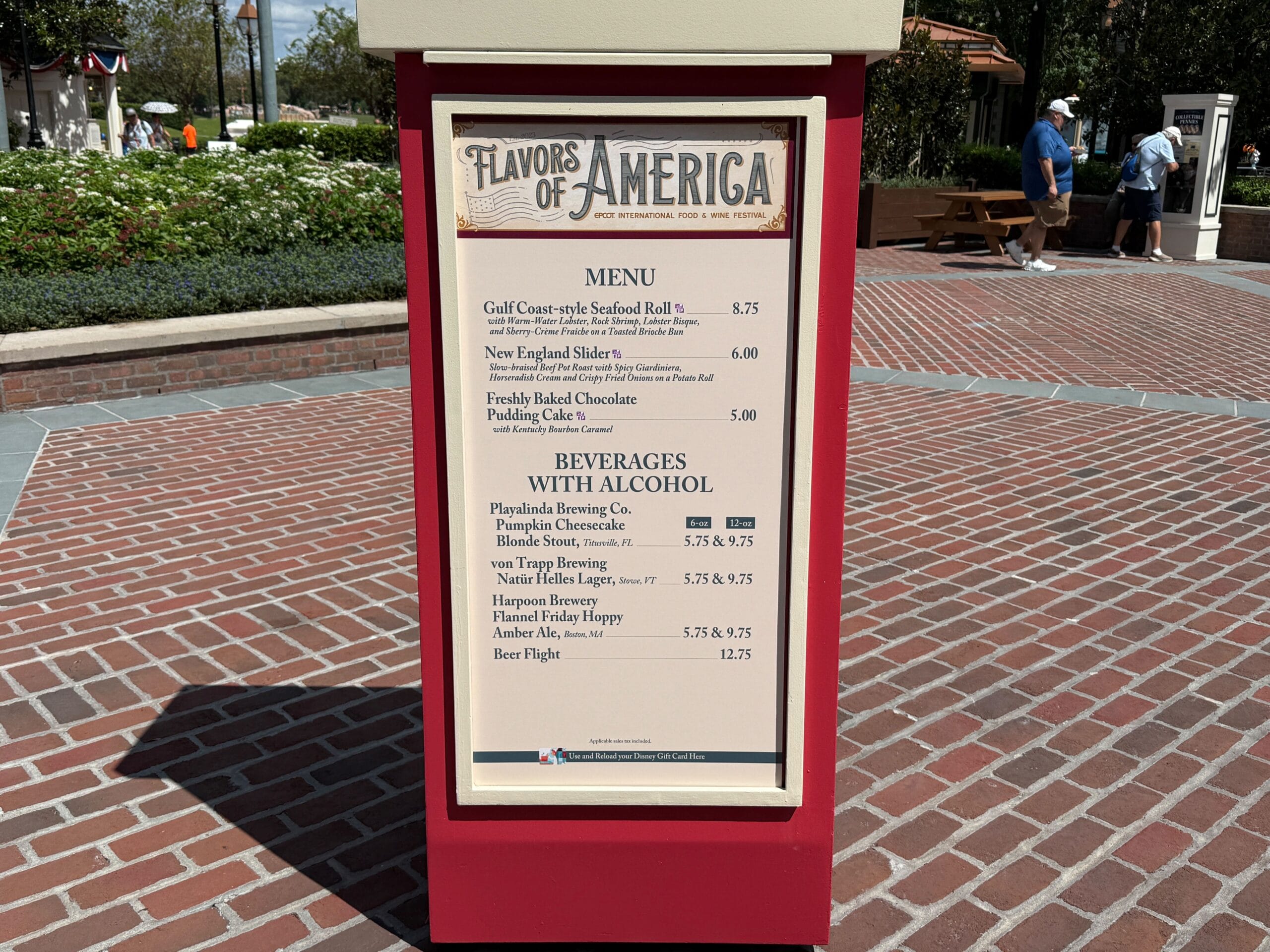 Menu board titled "Flavors of America" showcases Food & Wine with seafood, desserts, and cocktails on a brick walkway outside.
