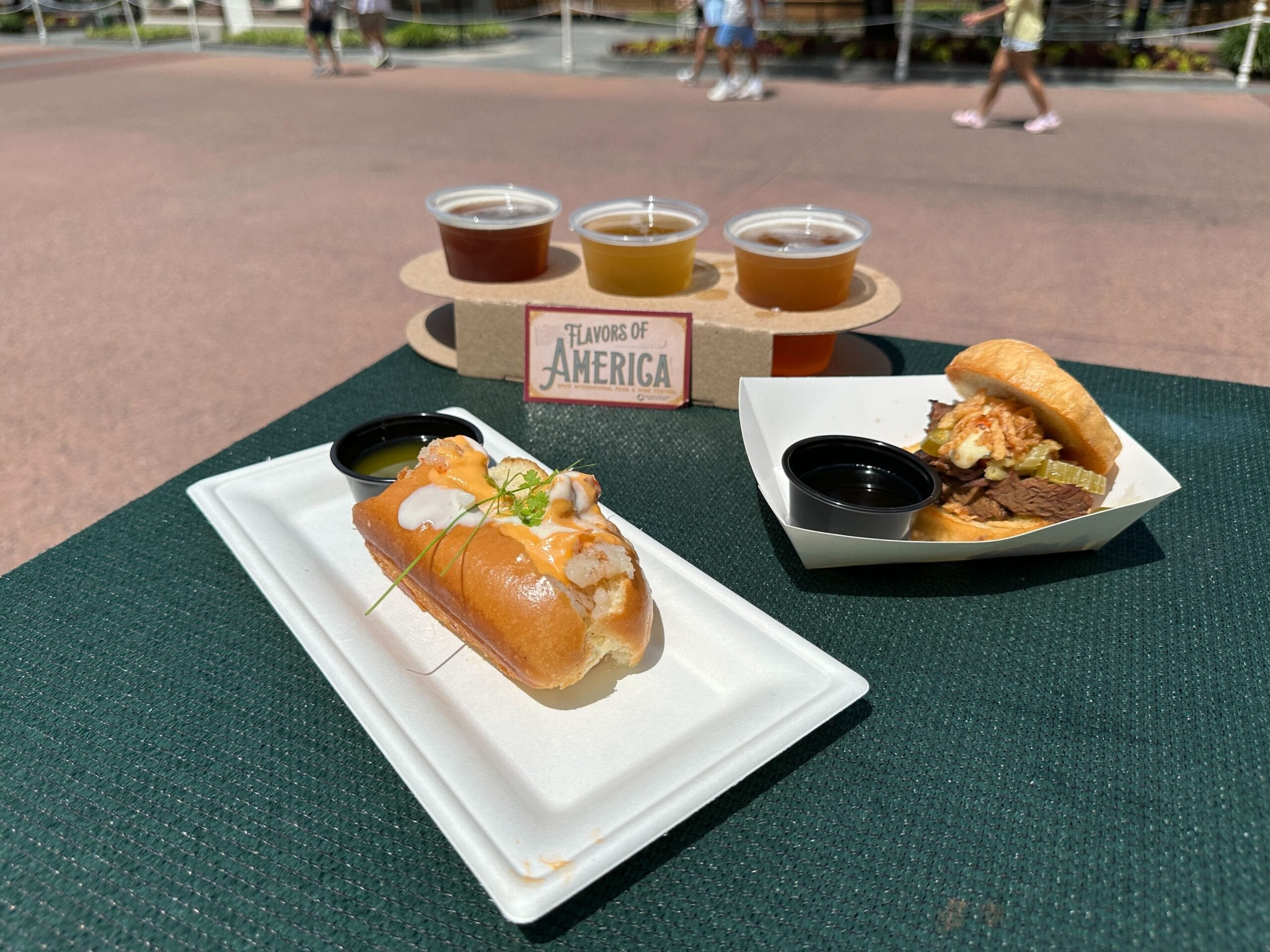 A lobster roll, beef slider, and four beers on a green table at the EPCOT Food and Wine Festival, "Flavors of America.