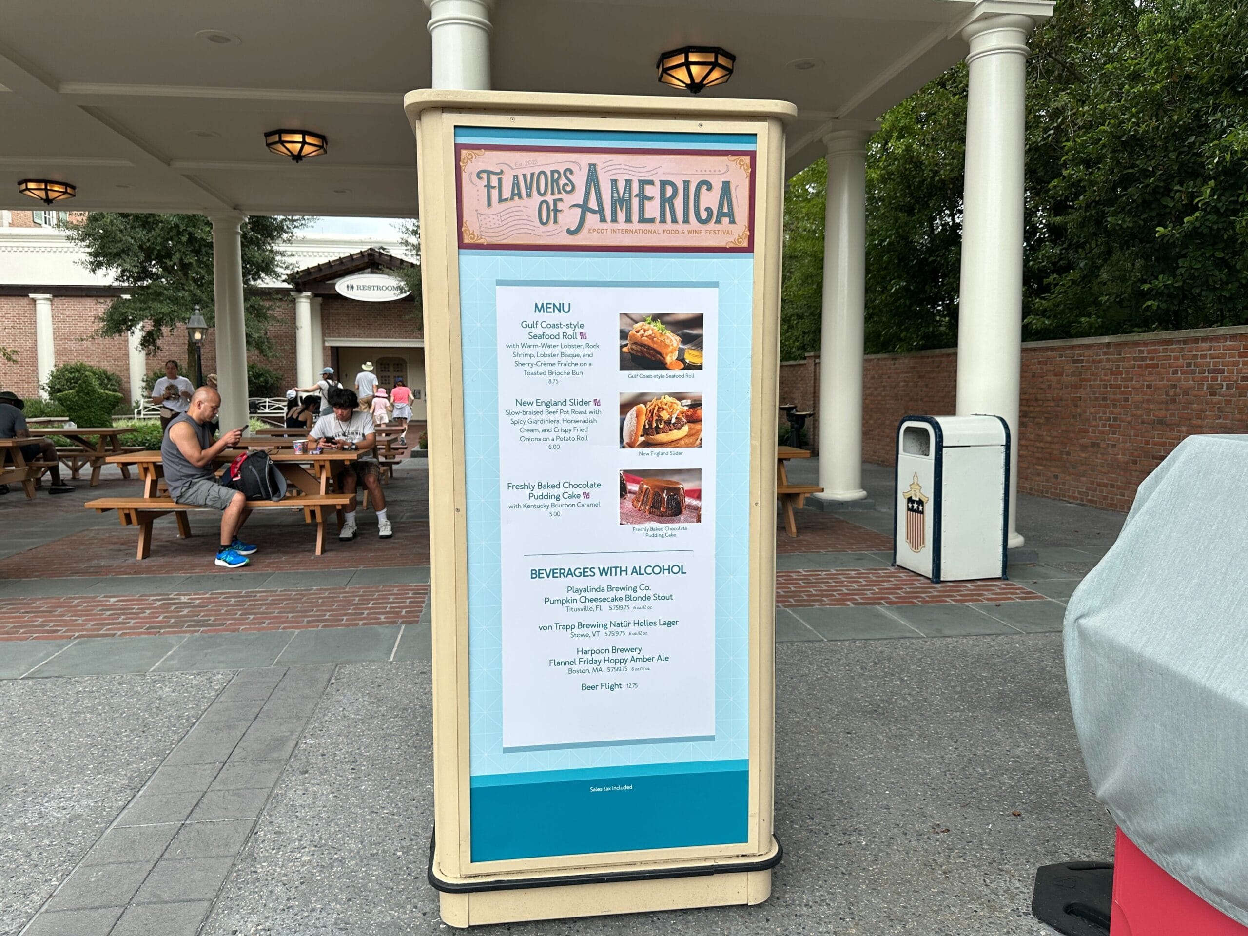 Flavors of America menu board outside features Food & Wine; people relax at picnic tables in the background.