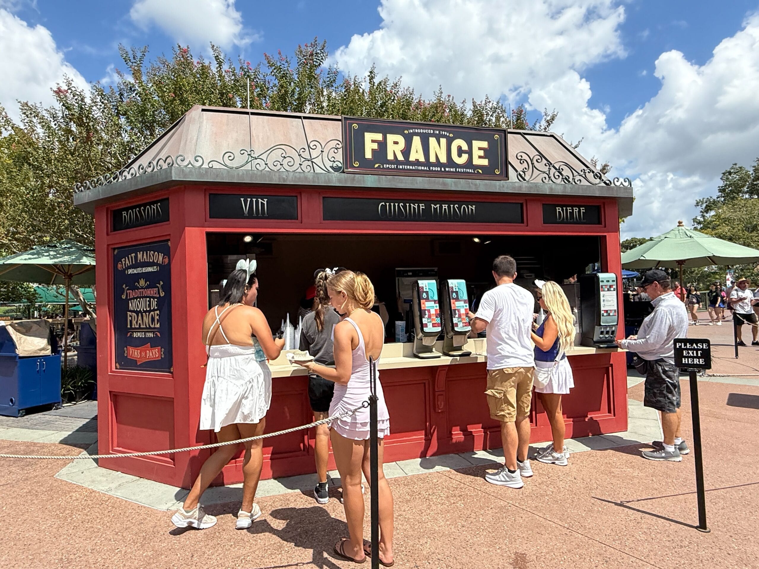 People order food & wine at a red France-themed outdoor booth under a sunny sky with trees and umbrellas in the background.
