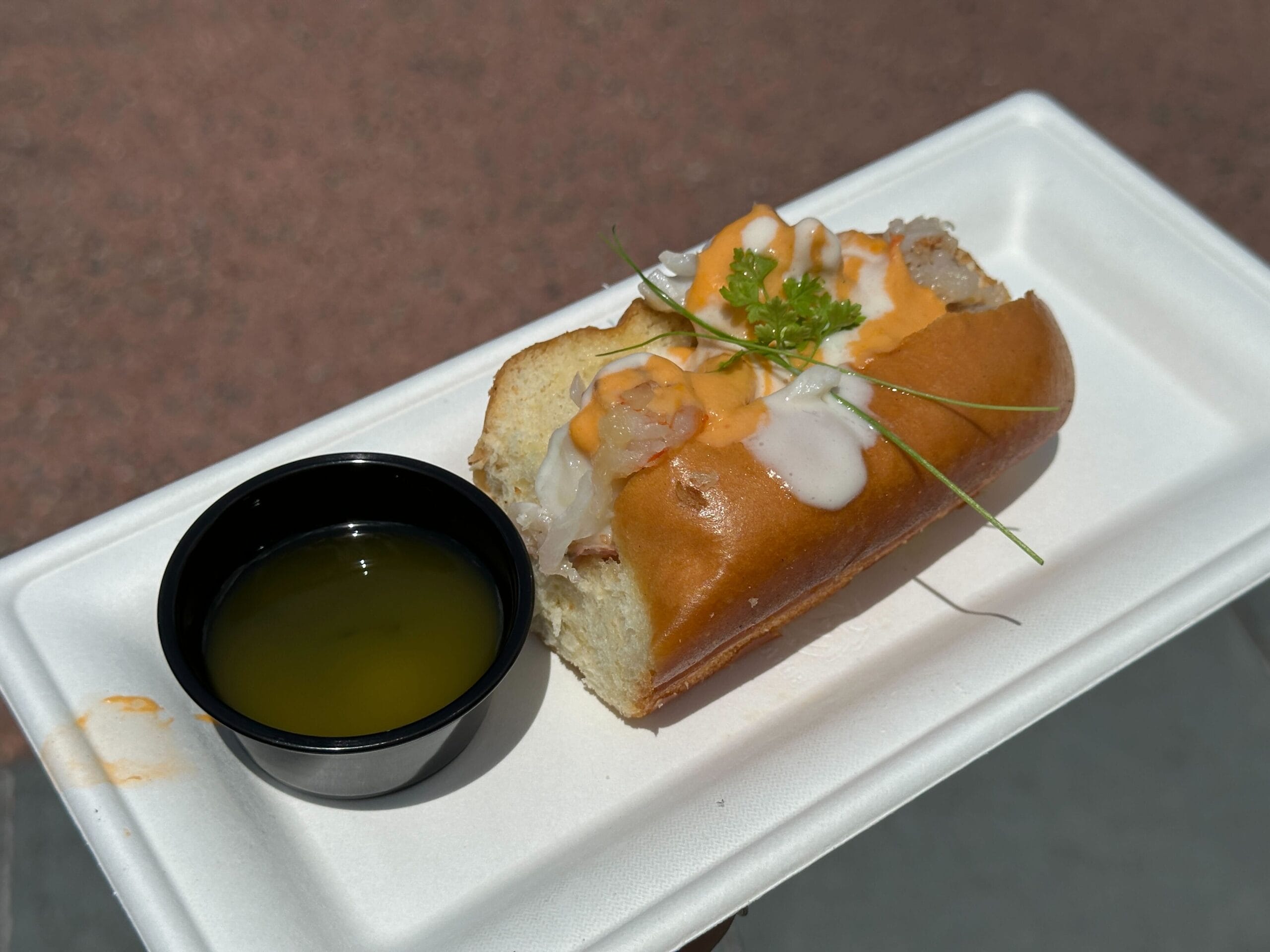 Lobster roll topped with sauce and herbs, Food & Wine style, served with melted butter on a white tray.
