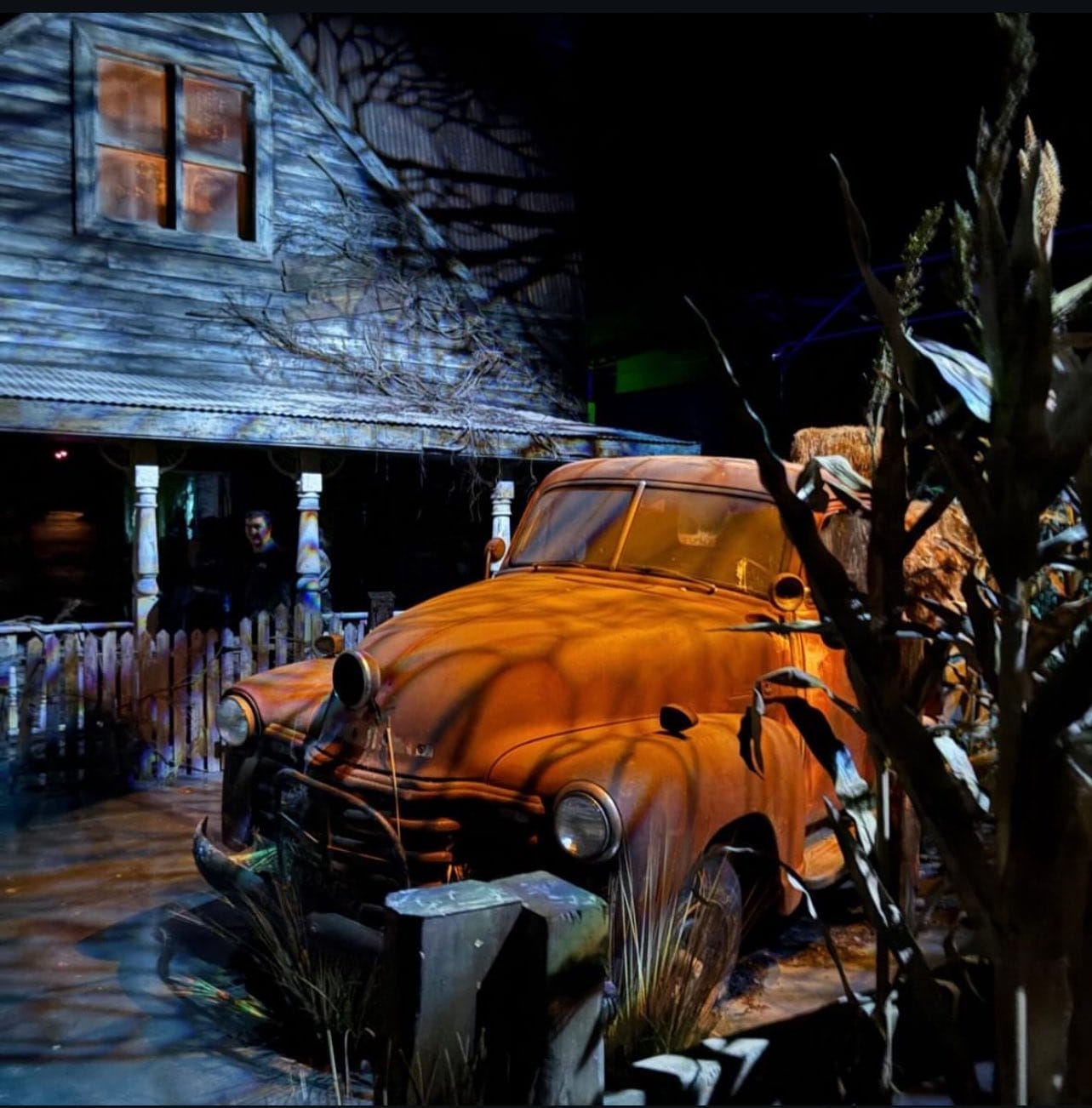 FIRST LOOK: A rusty old car sits before a spooky, dilapidated house at night, surrounded by eerie shadows and overgrown plants.