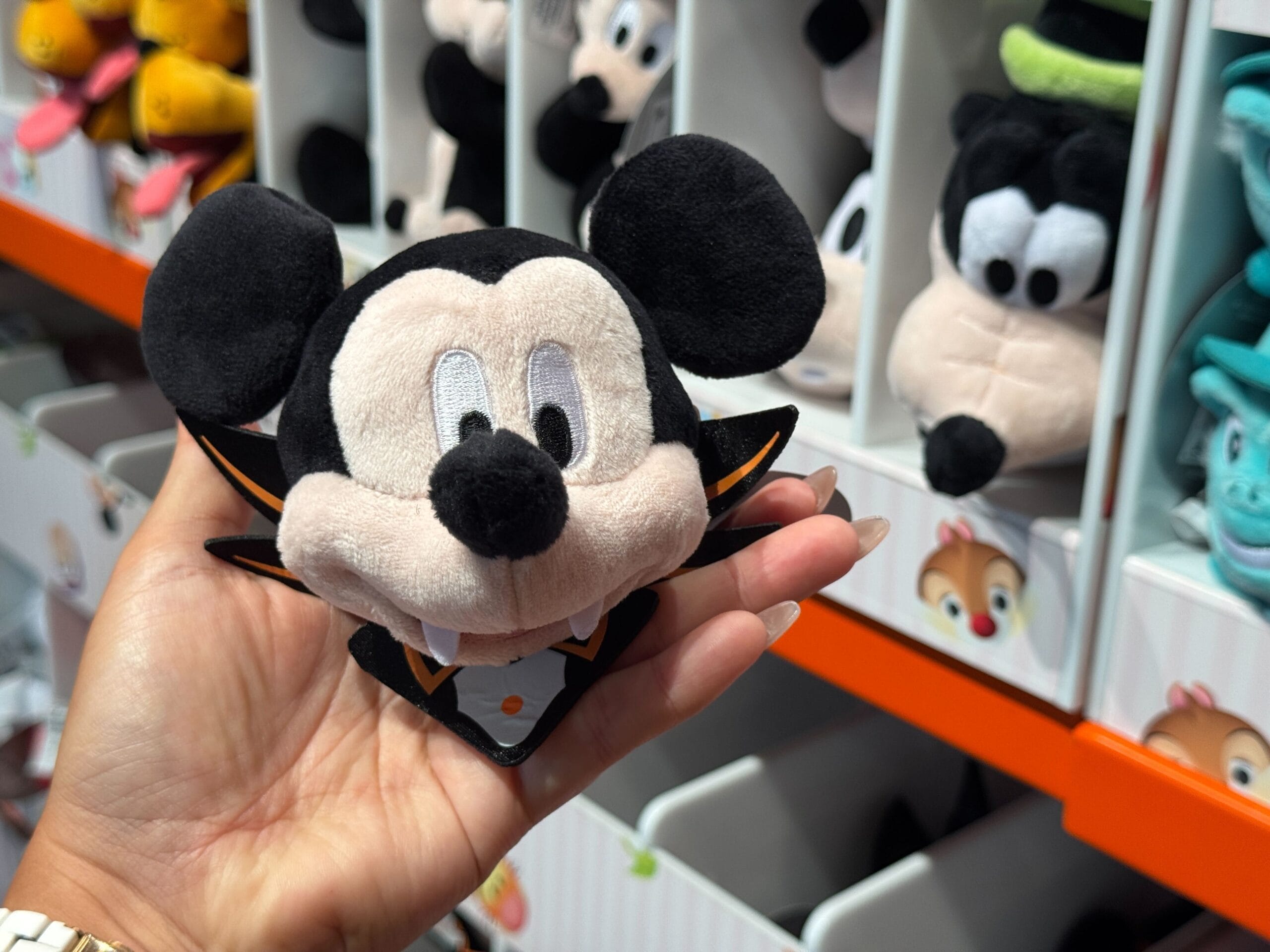 A hand holds a Mickey Mouse vampire plush, part of Walt Disney World's Halloween merchandise on shelves behind.