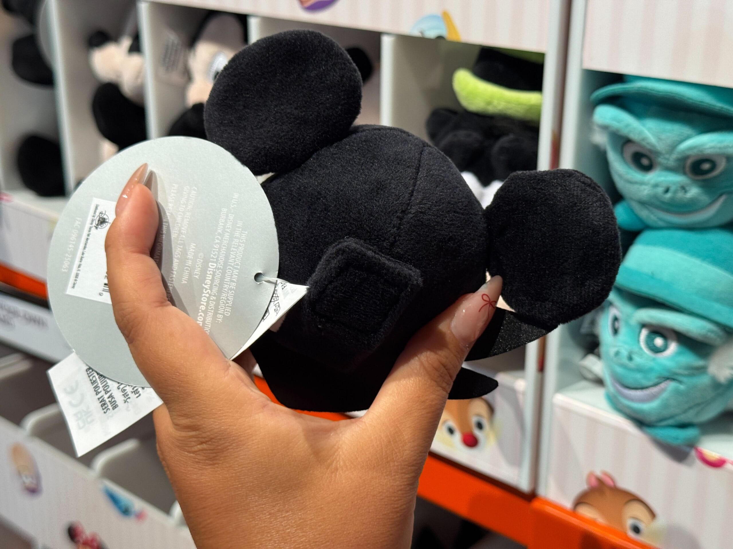 A hand holds a small black Mickey Mouse plush, part of Walt Disney World Halloween Merchandise, in front of stuffed toys.