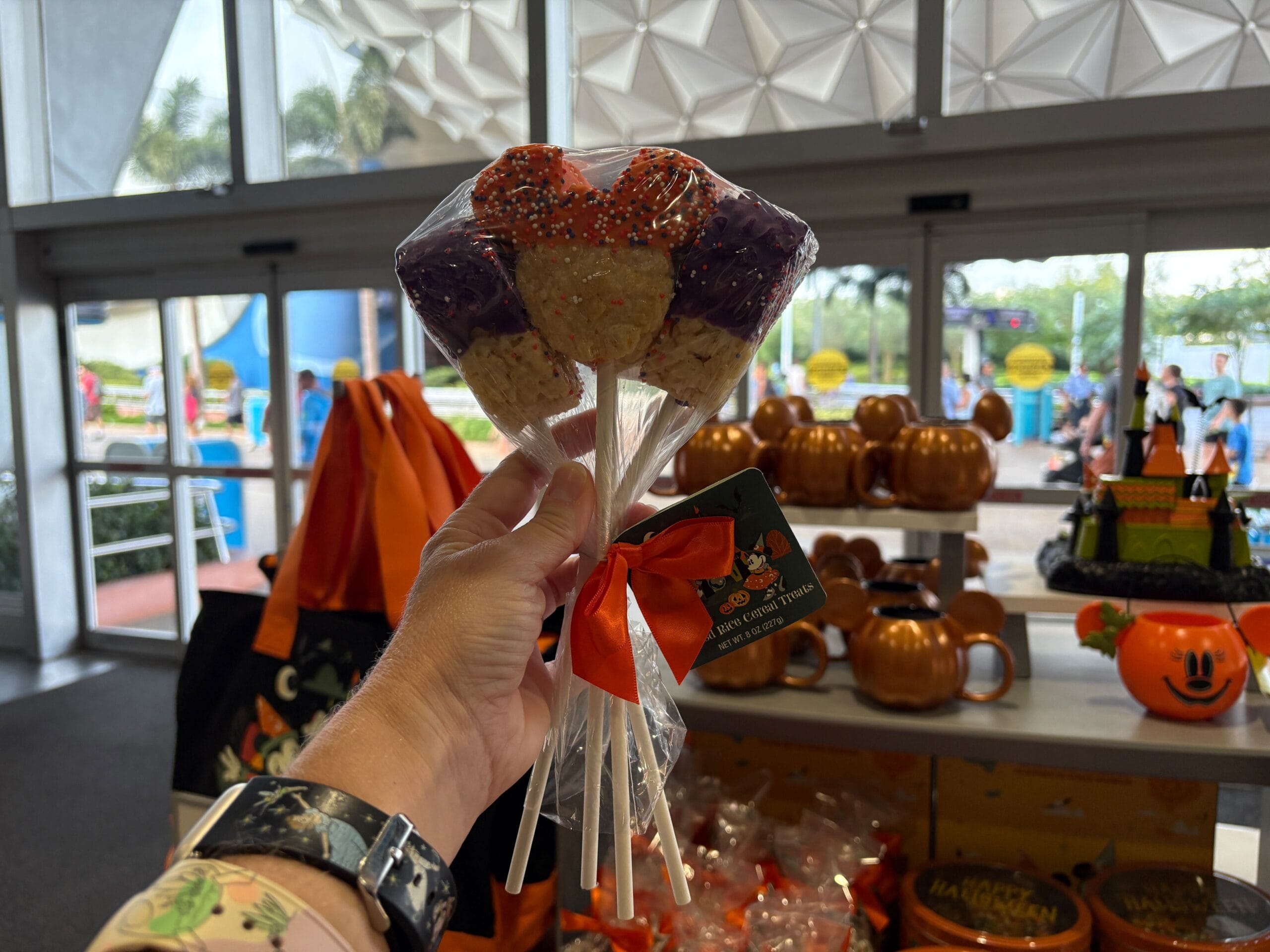 A hand holds three Halloween-themed Mickey treats inside a store filled with Disney Halloween Merch.