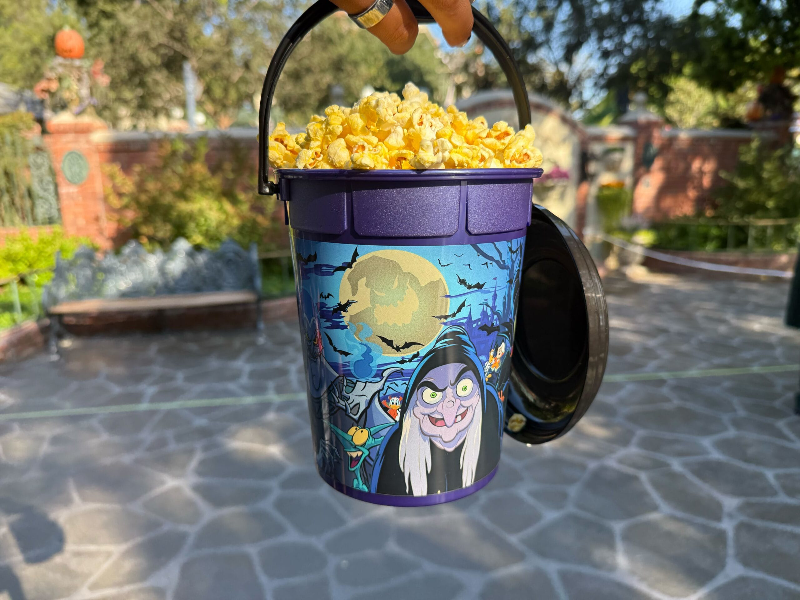 A hand holds a Villains-themed Halloween Popcorn Bucket with a witch design in an outdoor park at Disneyland.