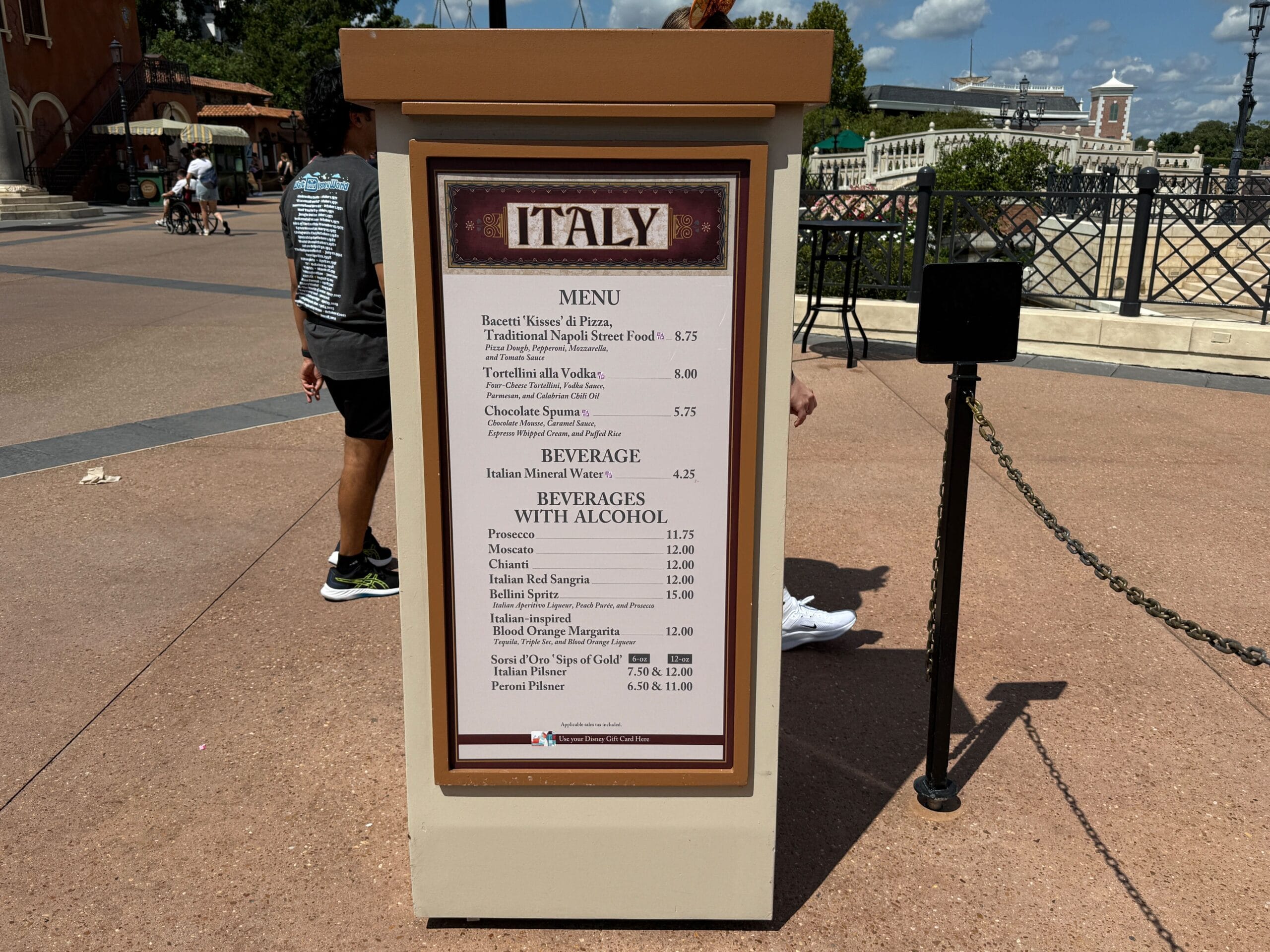 A New Food & Wine sign in Italy at Epcot lists menu items and beverage prices on a sunlit paved path.
