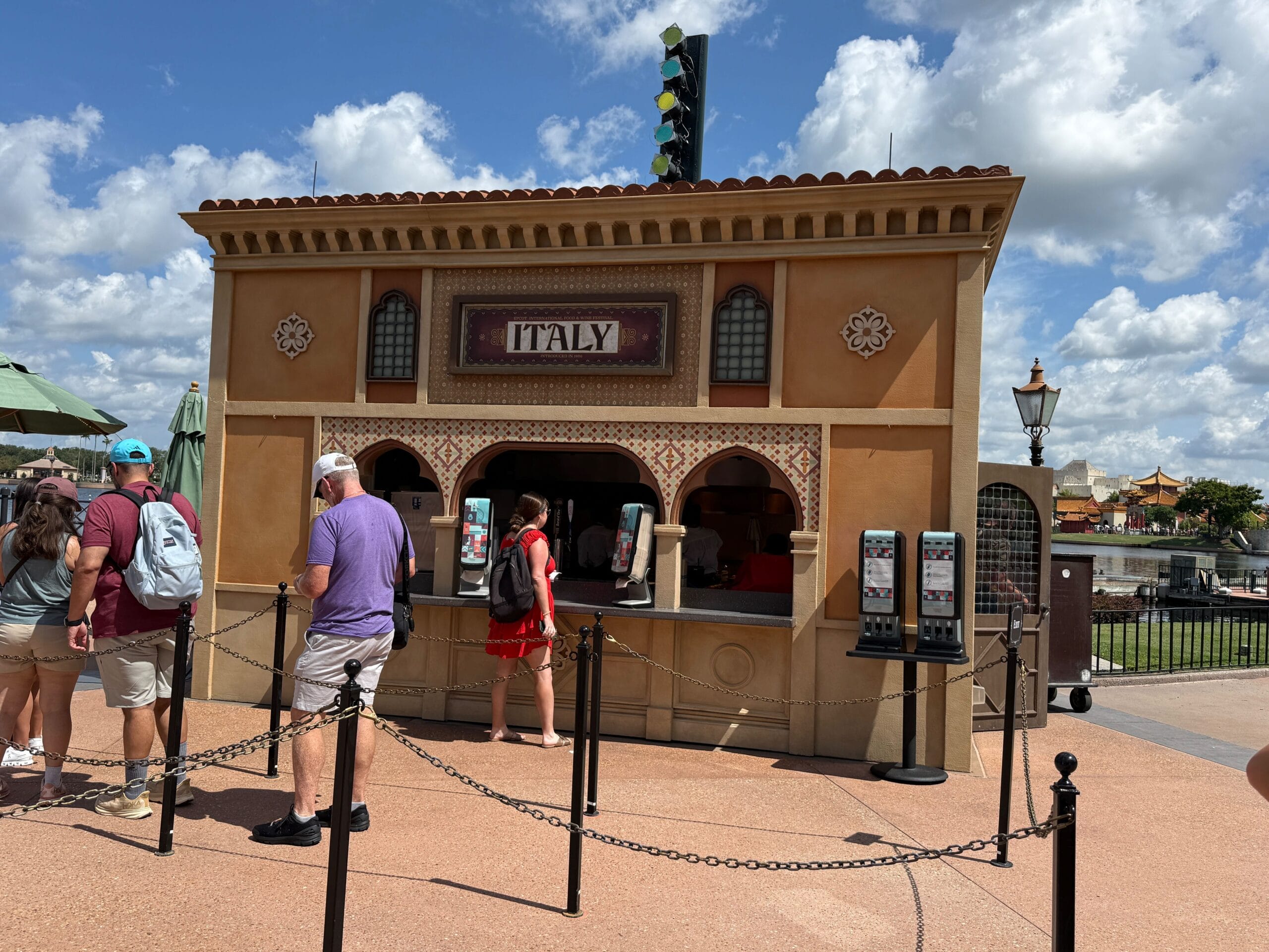 People stand in line at an outdoor Italy-themed food kiosk, eager to try New Food & Wine under a sunny sky.