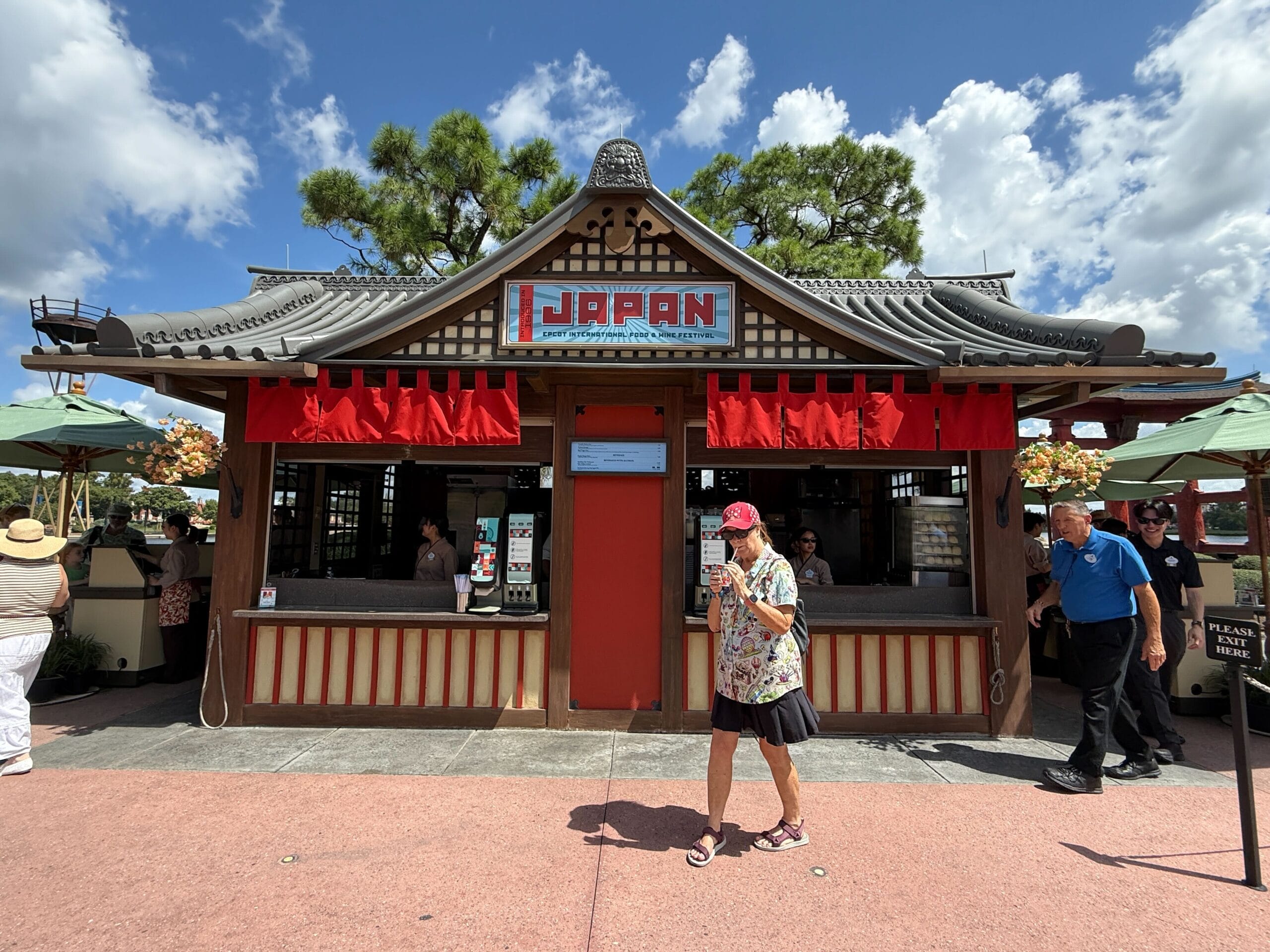 A person walks past a Food & Wine booth with a red sign and traditional roof under a bright, partly cloudy sky.