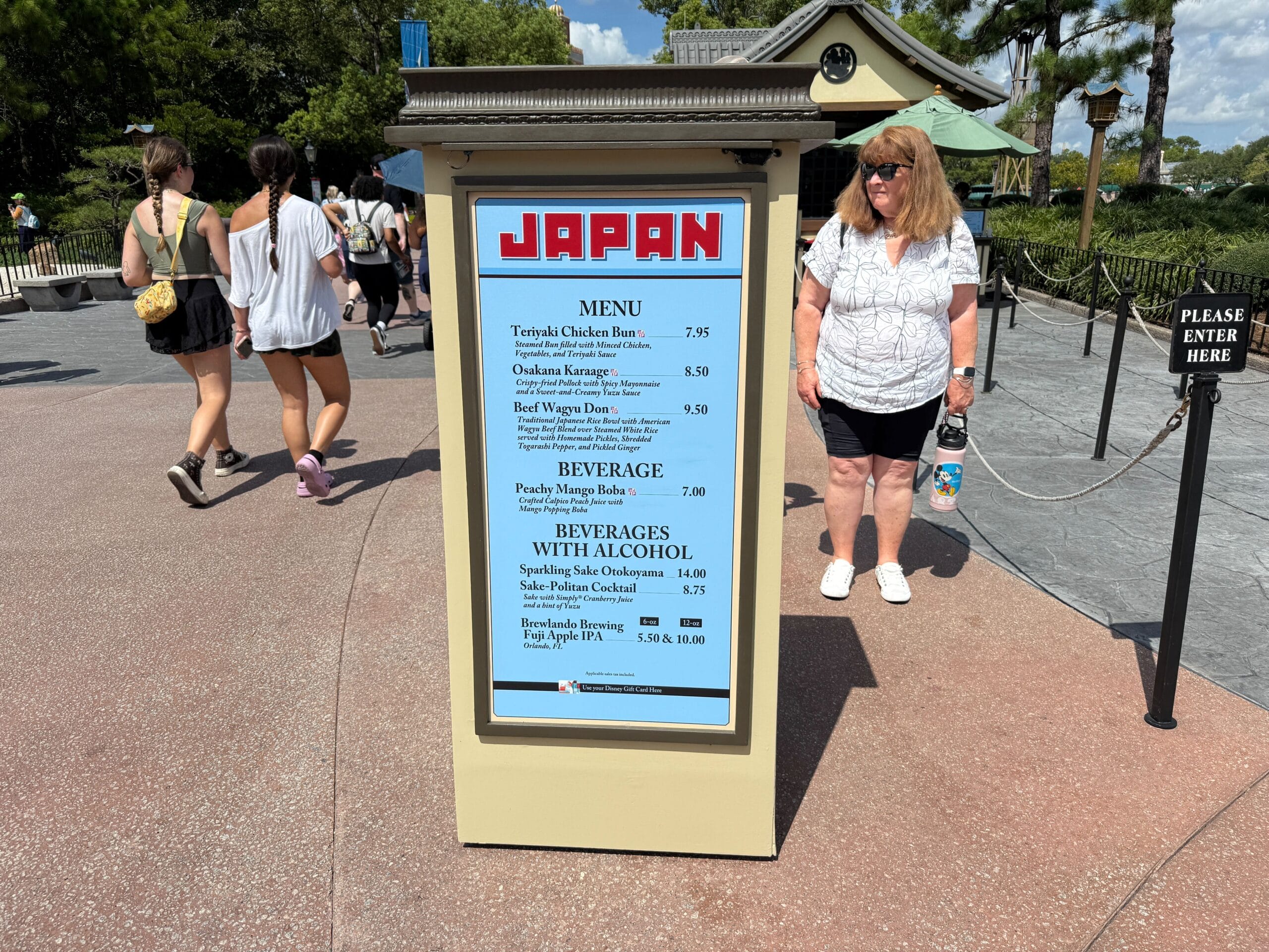 Outdoor Japan Food & Wine menu sign at EPCOT, listing items, with people strolling by on a sunny day.