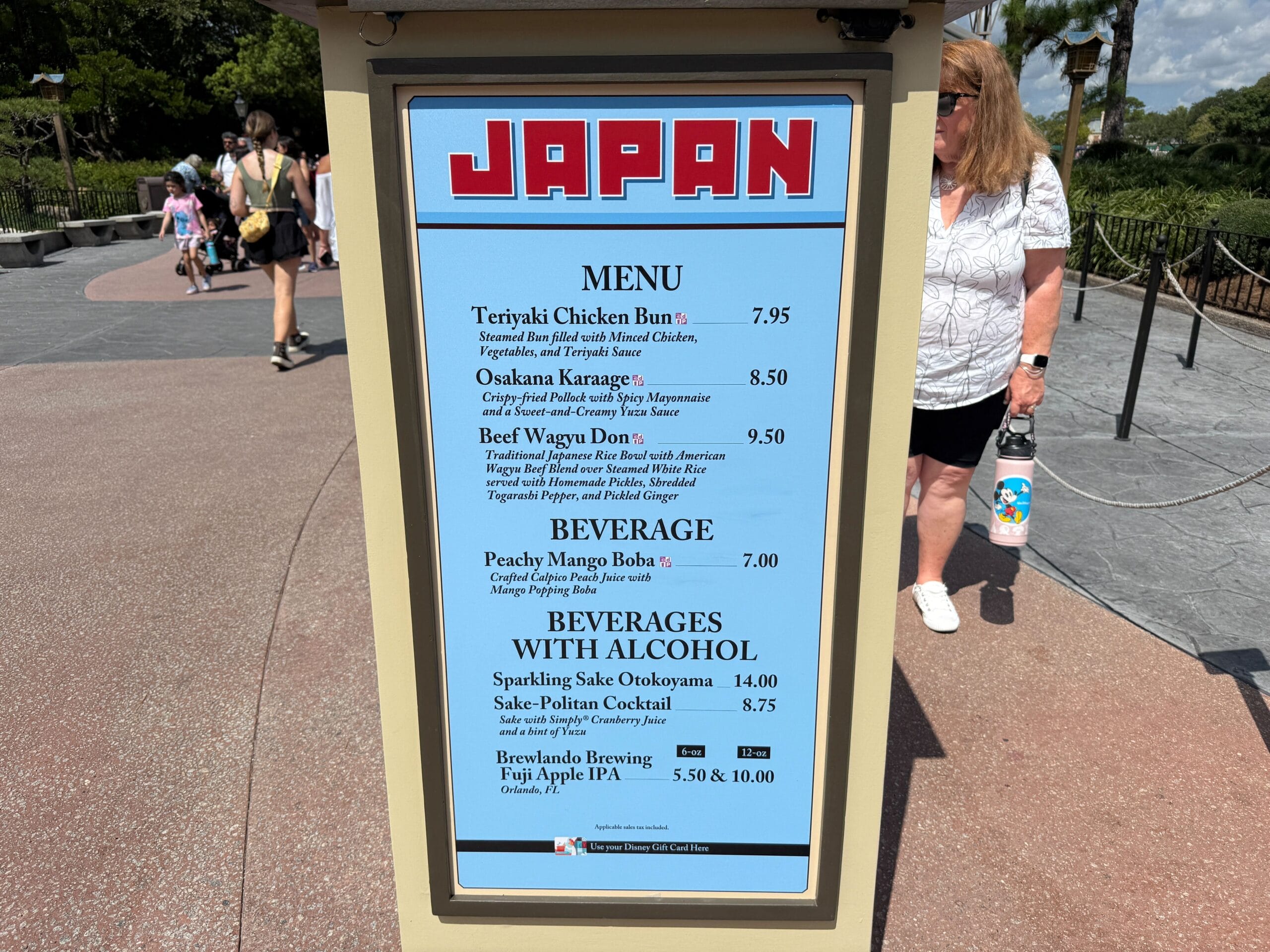 Menu sign for the Japan pavilion at Food & Wine, listing food, beverages, and alcoholic drinks; people and trees in background.