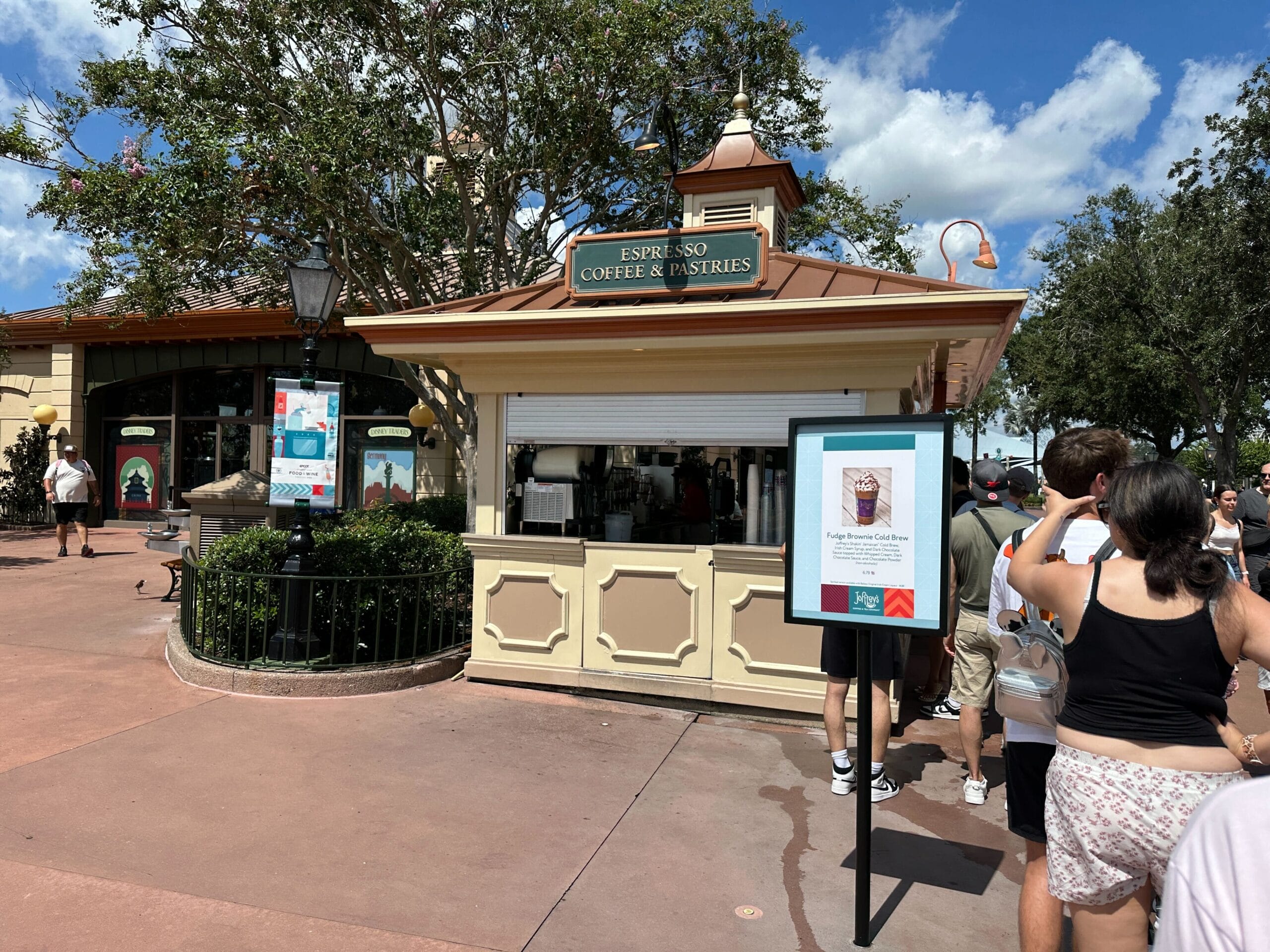 People wait in line at a New Food & Wine outdoor espresso, coffee, and pastries kiosk on a sunny day with trees behind.