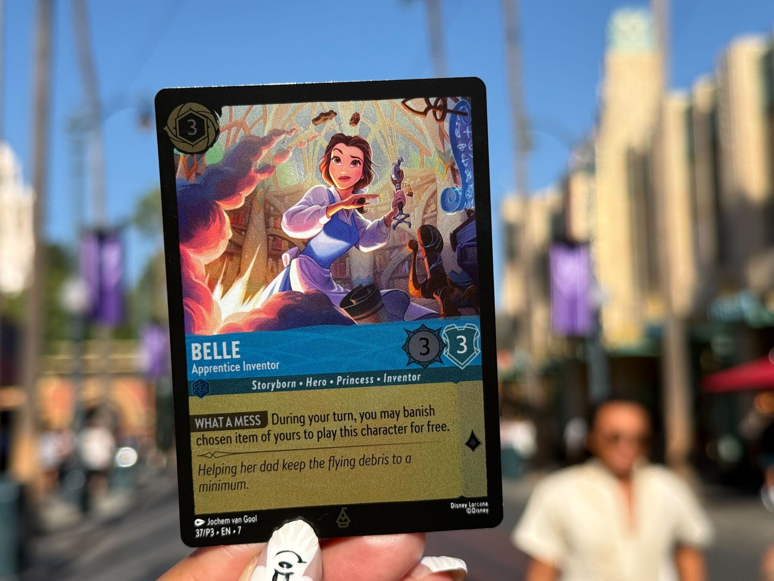 A hand holds a Lorcana Card of Belle in front of a sunny street scene near Disney Parks, with blurred people and buildings.