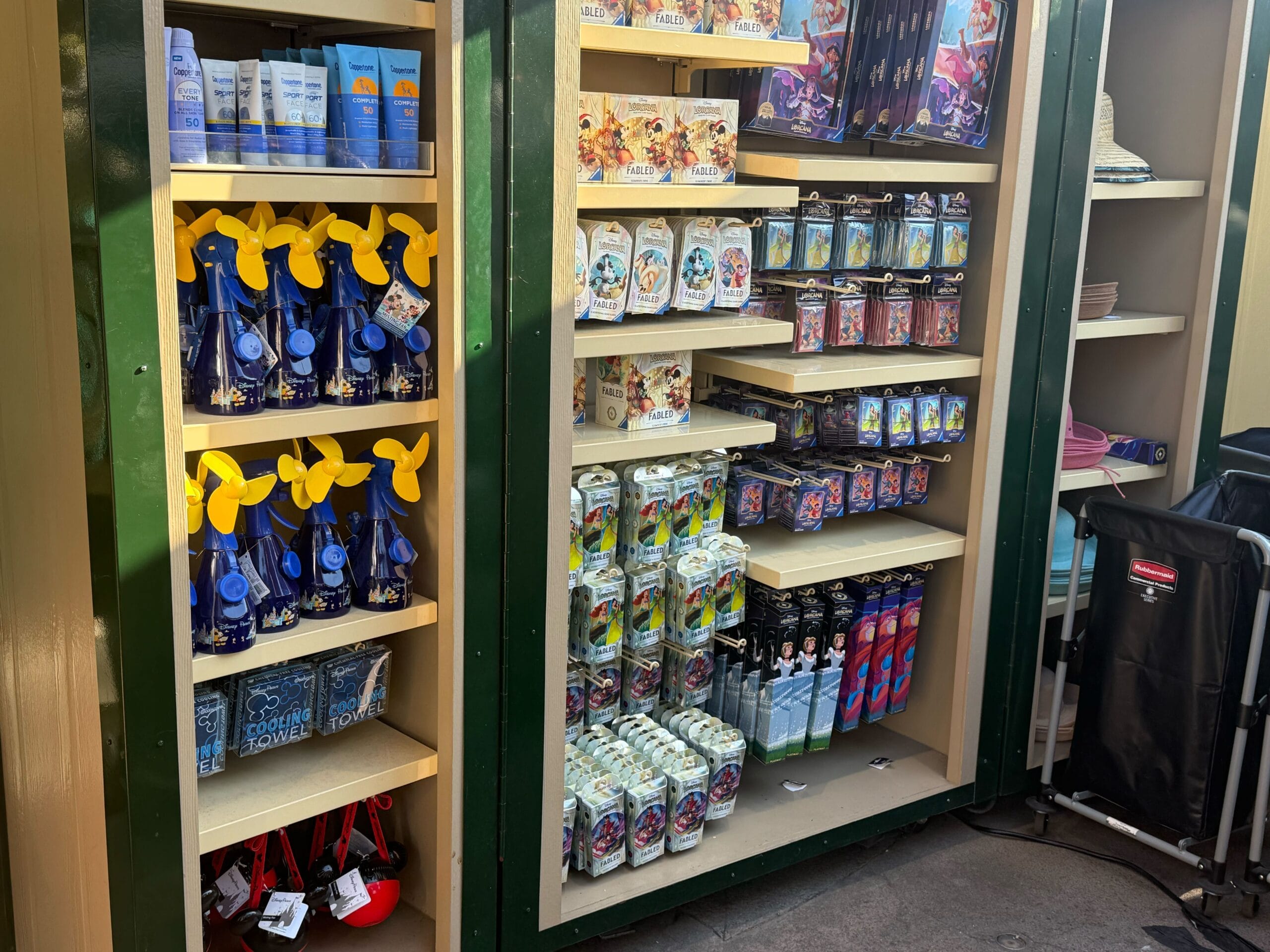 Shelves display water fans, sunscreen, Lorcana Card packs, and drink bottles at an outdoor merchandise stand.