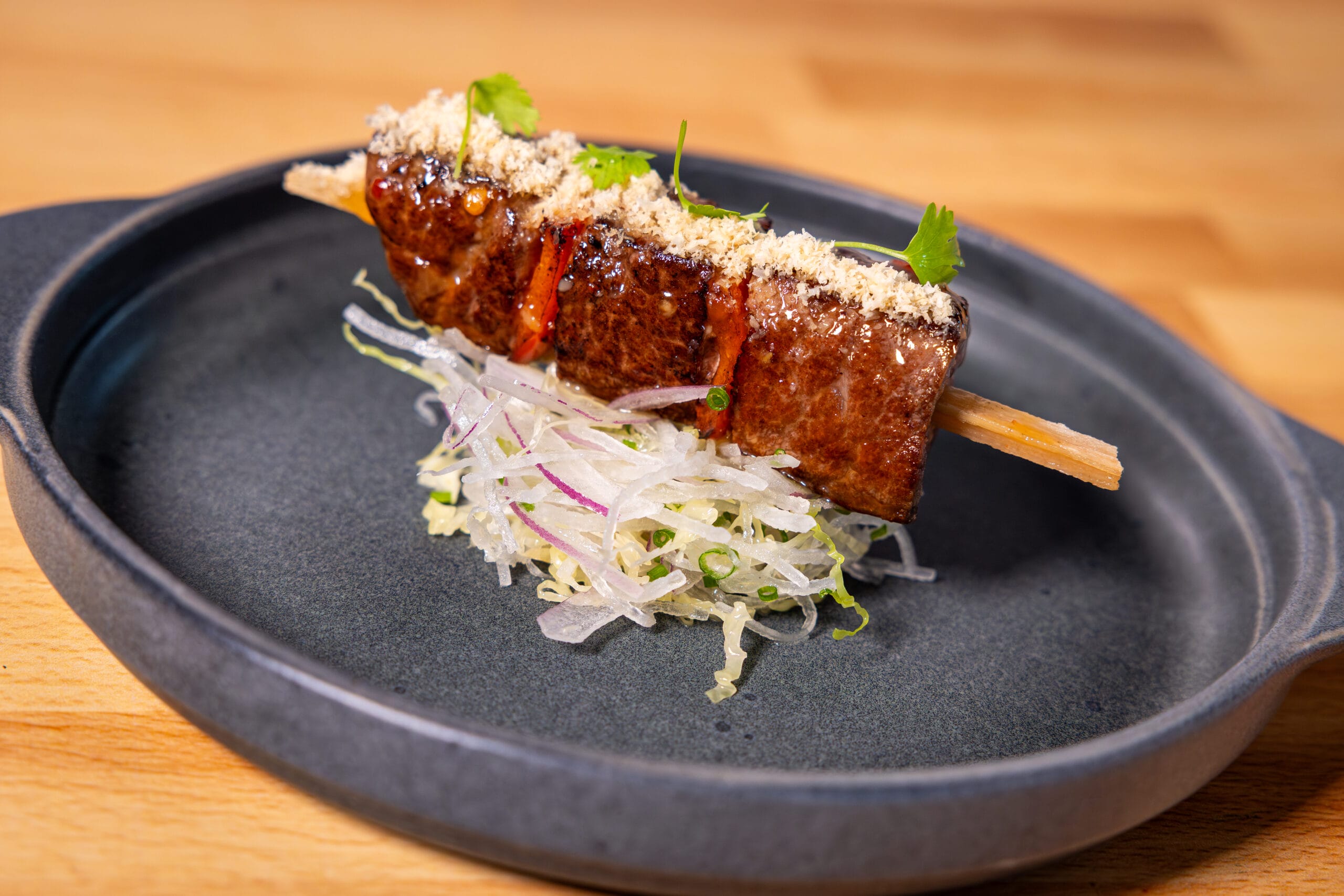 A grilled meat skewer garnished with herbs and sauce, served over shredded vegetables on a black plate.
