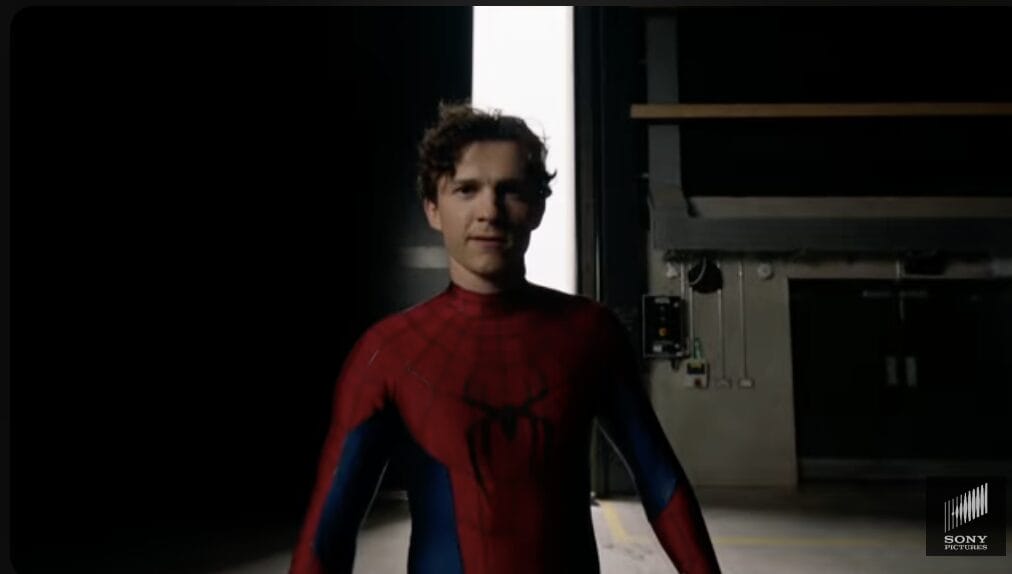 A person in a Tom Holland Spider-Man suit stands indoors, light from an open doorway creating a teaser vibe.