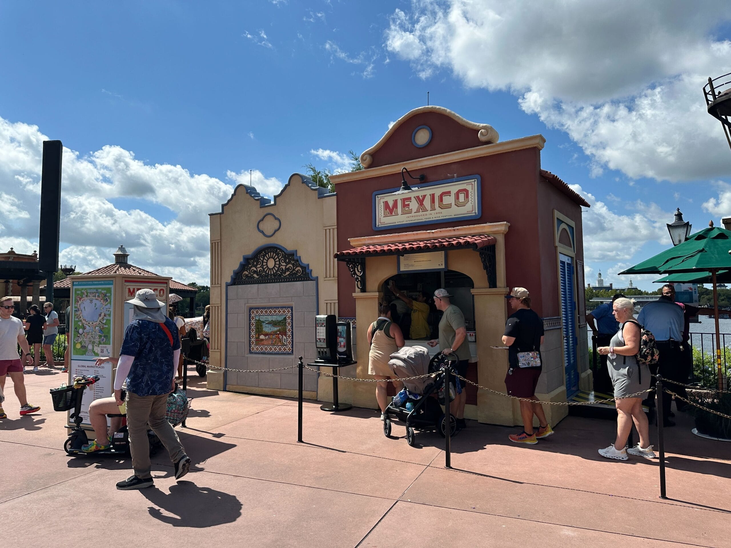 People stand in line outside the colorful Mexico pavilion, eager for Food & Wine delights under the blue theme park sky.