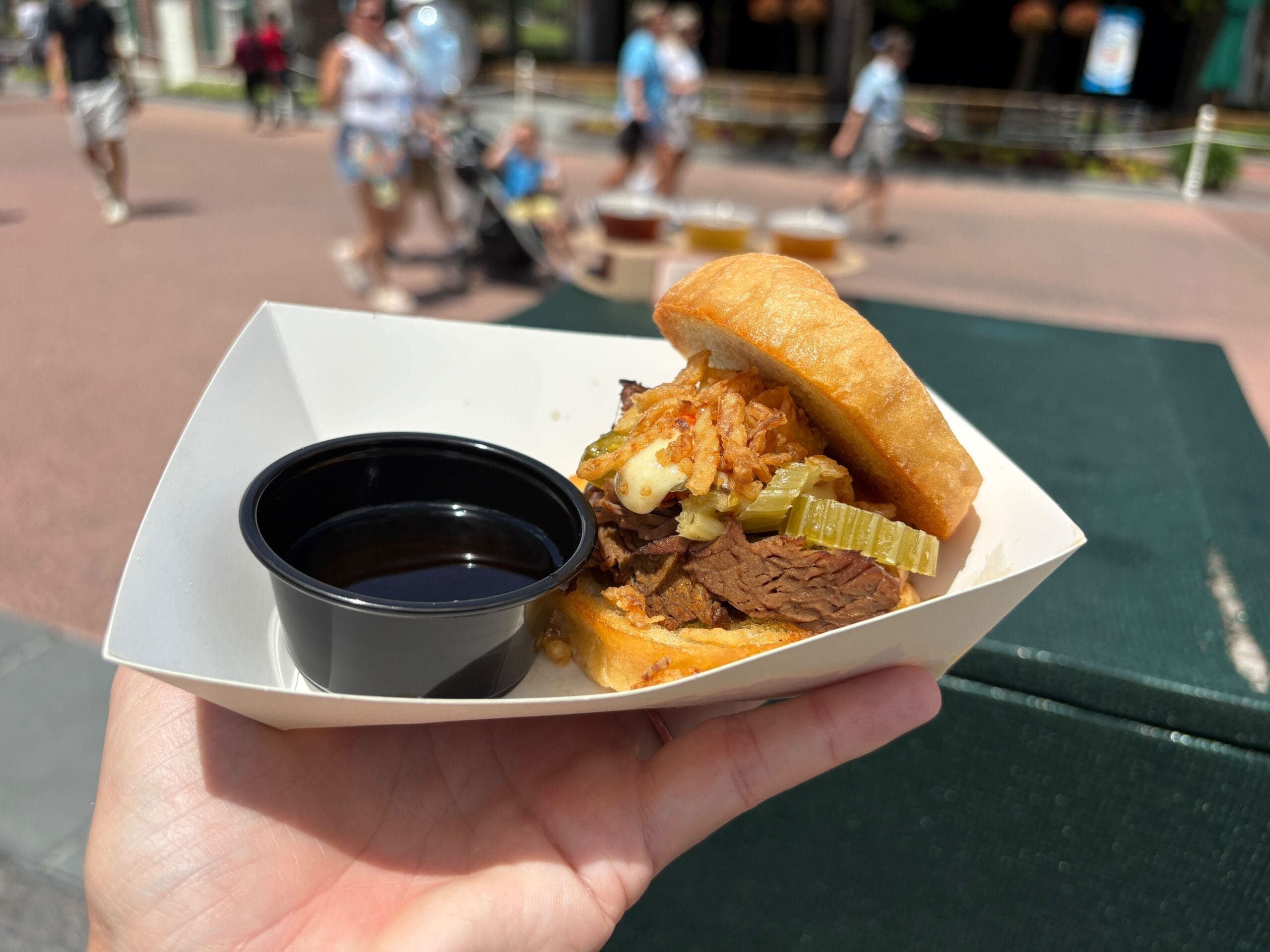 A hand holds a beef sandwich with fried onions and dipping sauce—perfect for any Food & Wine outdoor gathering.