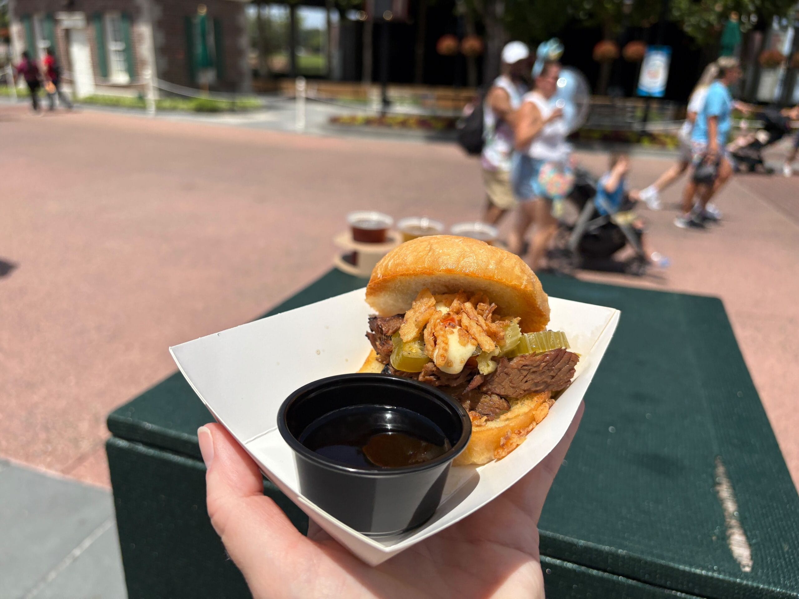 A hand holds a small beef sandwich with fried onions and sauce, perfect for any Food & Wine lover; city life blurs behind.